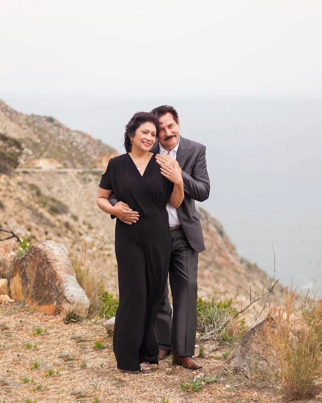 Older couple embracing on a rocky hillside above the sea, she in a black jumpsuit and he in a grey suit.