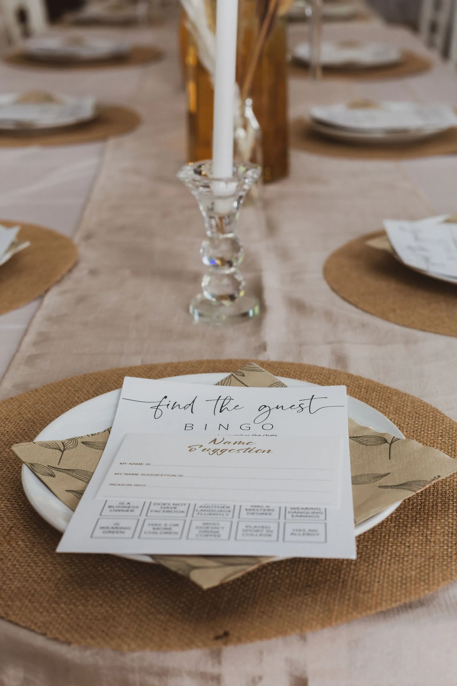Place setting with a Find the Guest bingo card and a baby name suggestion card on a white plate and jute mat, a crystal candlestick behind.