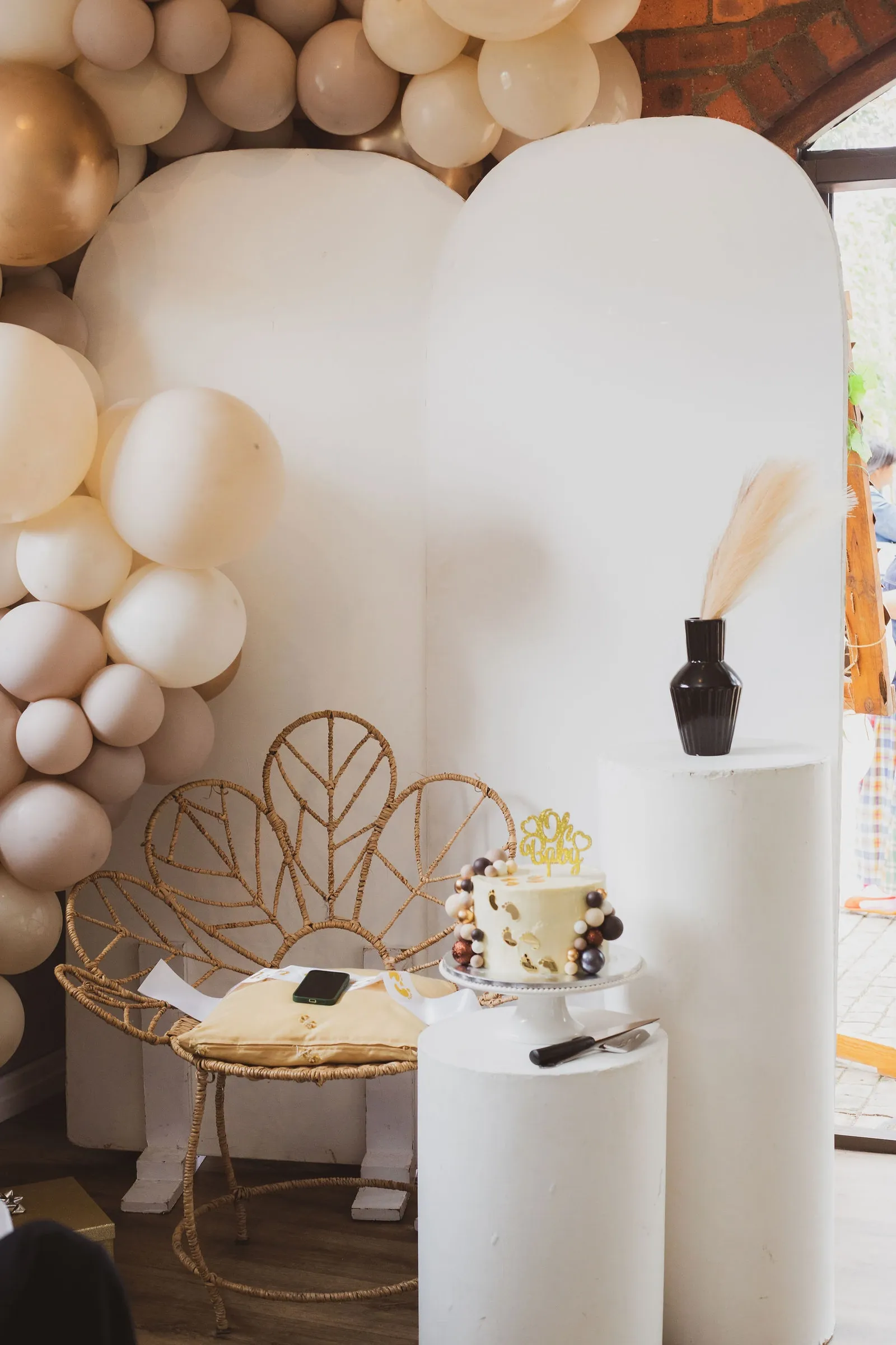 Cream, blush and gold balloon garland around two white arch boards, a rattan fan-back chair and an Oh Baby cake on a white plinth.