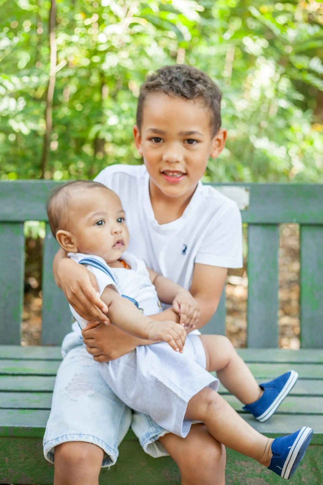 Older boy on a green garden bench holding his baby brother on his lap, the baby in blue shoes looking to the side.
