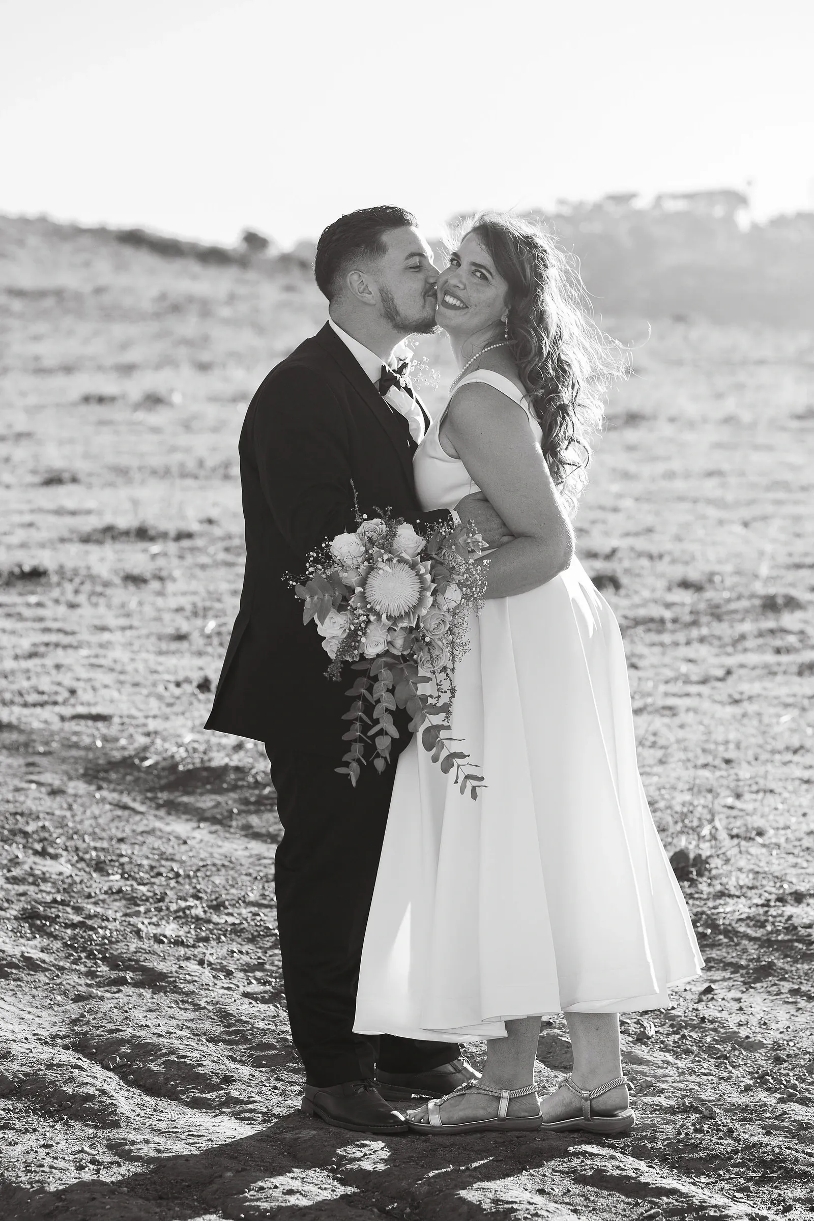 Black and white photo of a groom kissing his bride's cheek as she smiles at the camera, holding a bouquet with a protea and trailing eucalyptus.