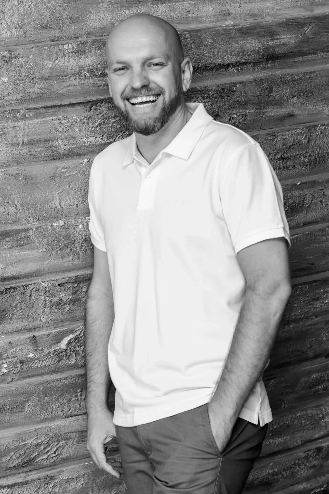 Black and white portrait of a laughing bearded man in a white polo shirt leaning against a textured wall.