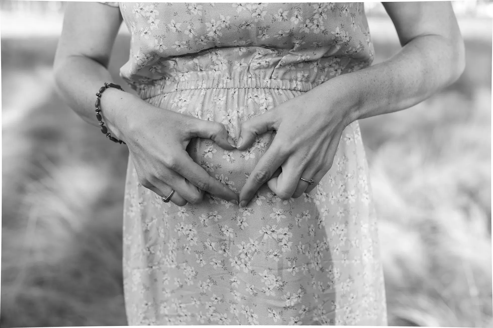 Black and white close-up of hands shaping a heart on a pregnant bump in a floral dress.