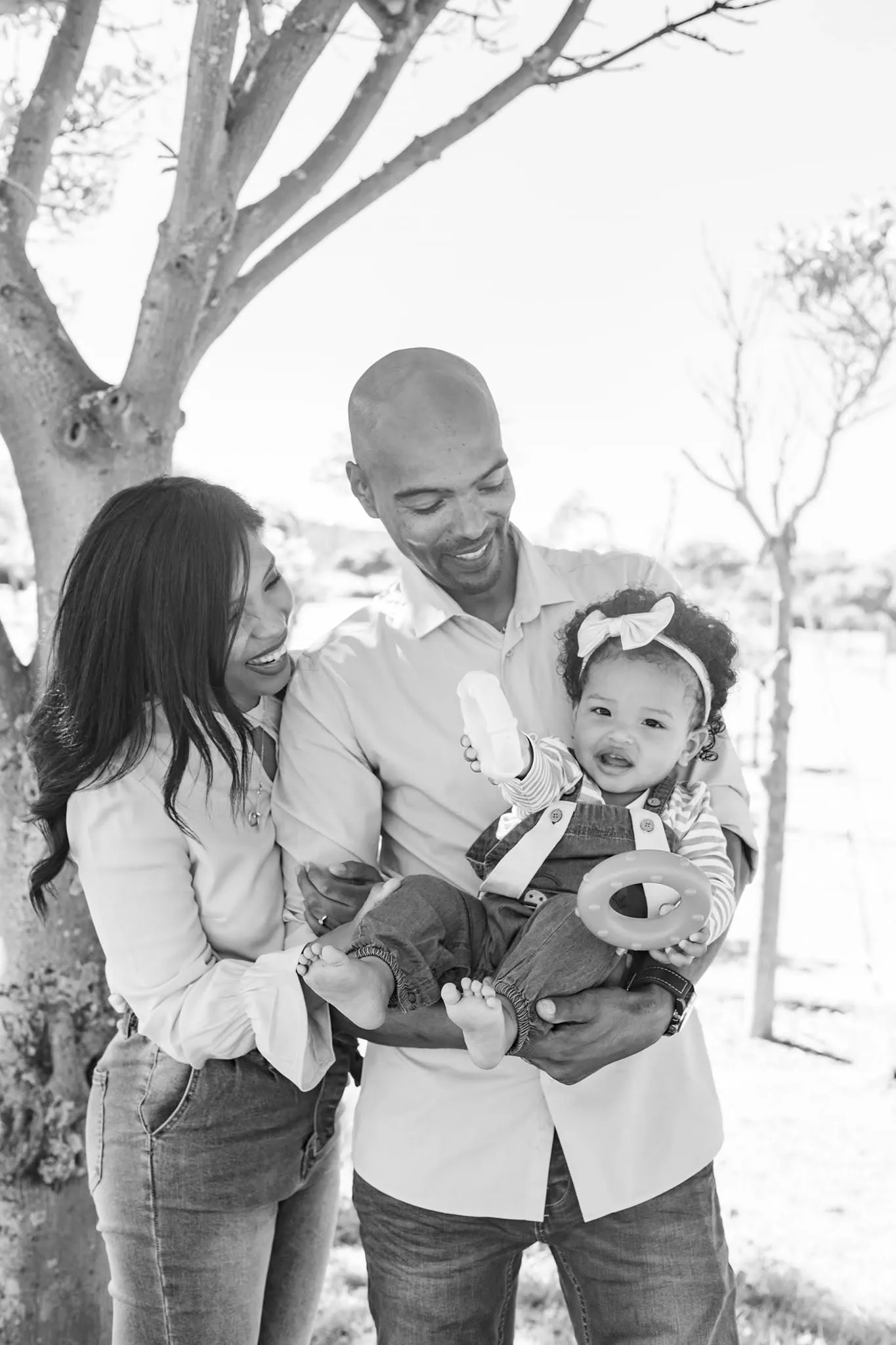 Black and white photo of parents laughing with their baby girl, who wears a bow headband and holds a toy ring.
