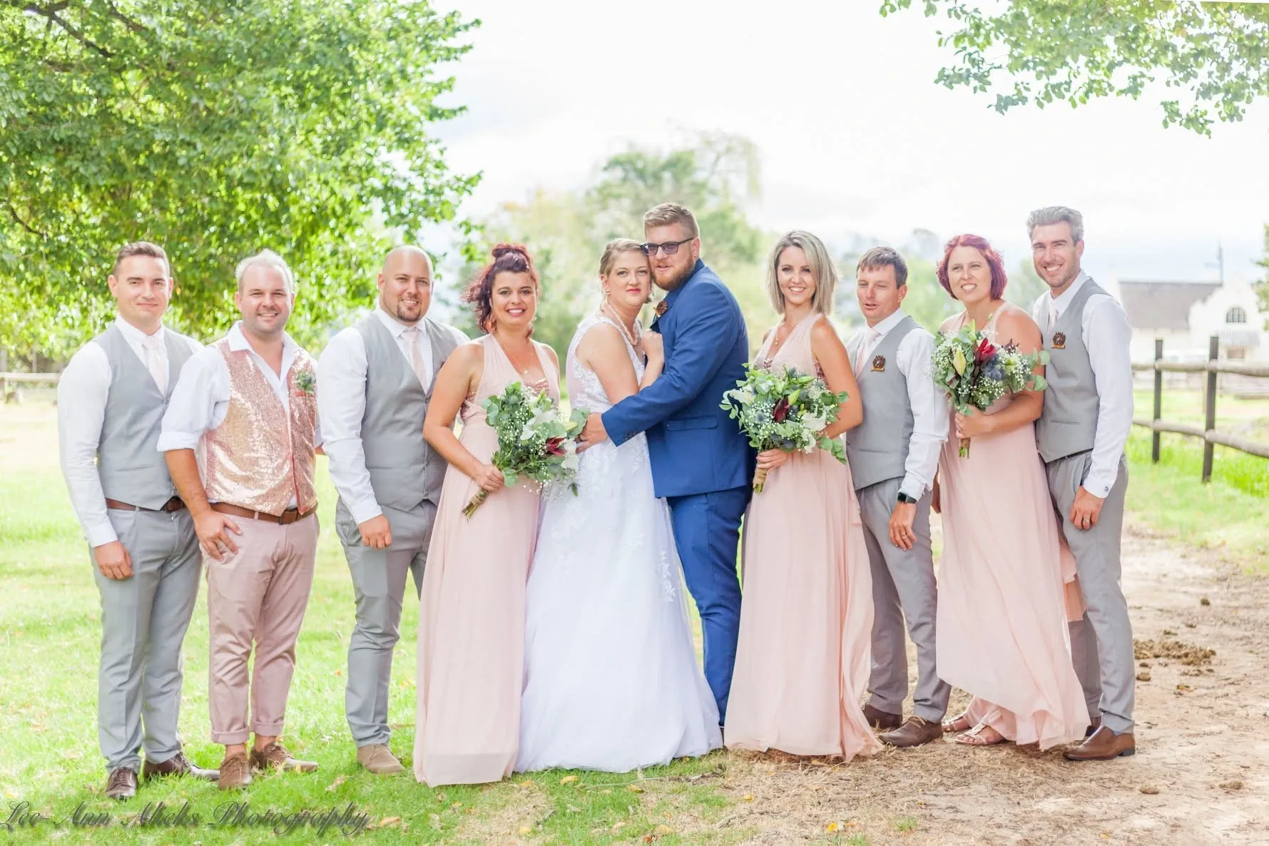 Bride and groom in the middle of their bridal party, bridesmaids in blush dresses and groomsmen in grey waistcoats, on a farm lawn.