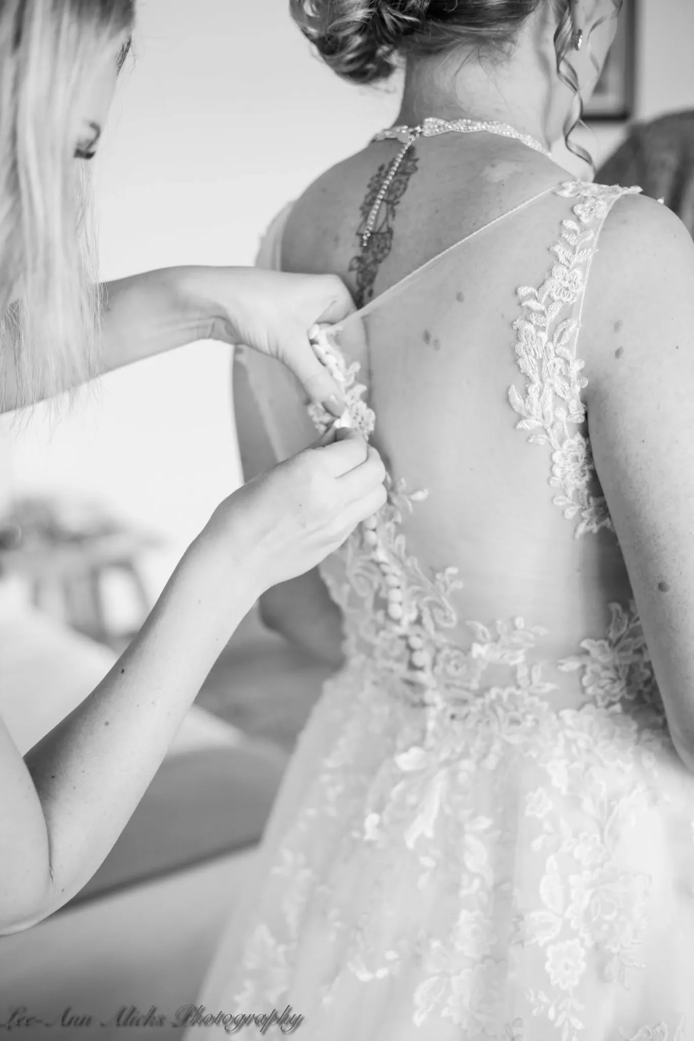 Black and white close-up of hands fastening the covered buttons on the sheer lace back of a wedding gown.