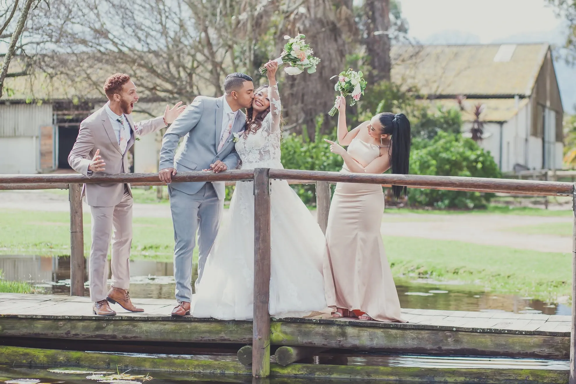 Bride and groom kissing on a wooden footbridge while a groomsman and a bridesmaid cheer, the bride's bouquet raised high.