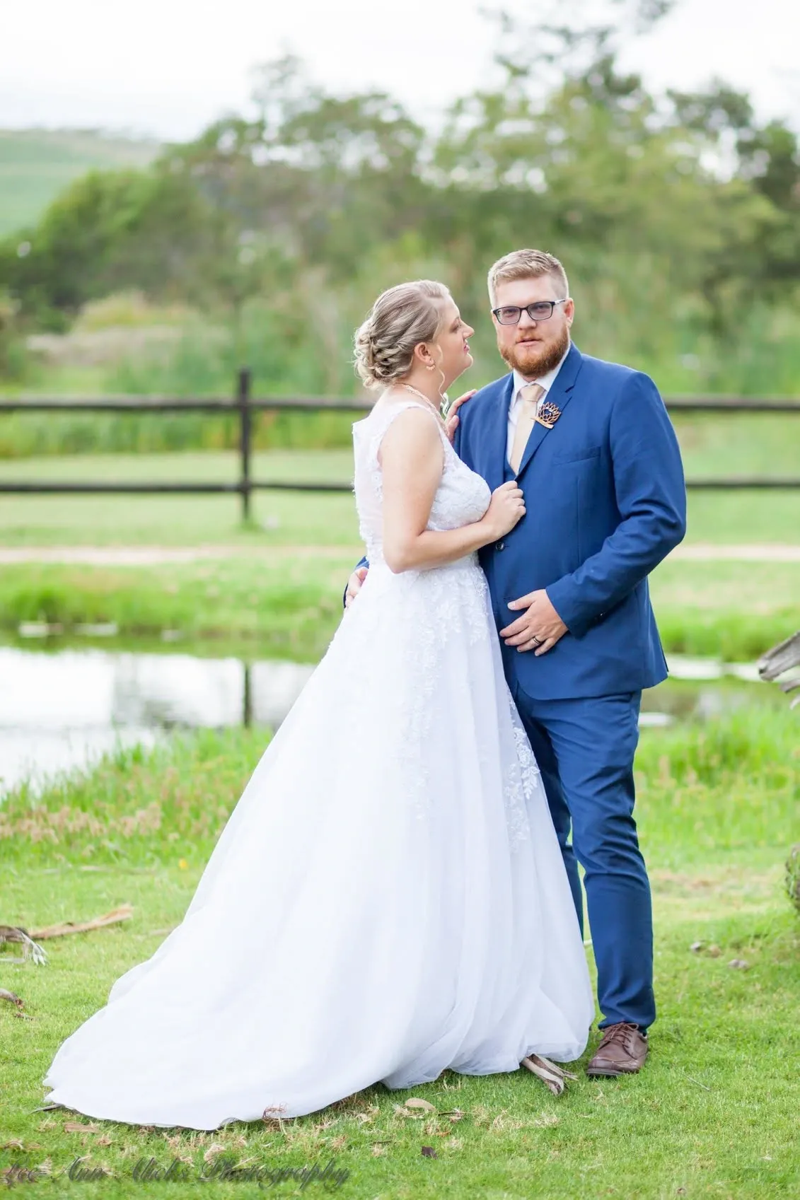 Bride in a lace gown and groom in a blue suit standing close beside a pond and a wooden farm fence.