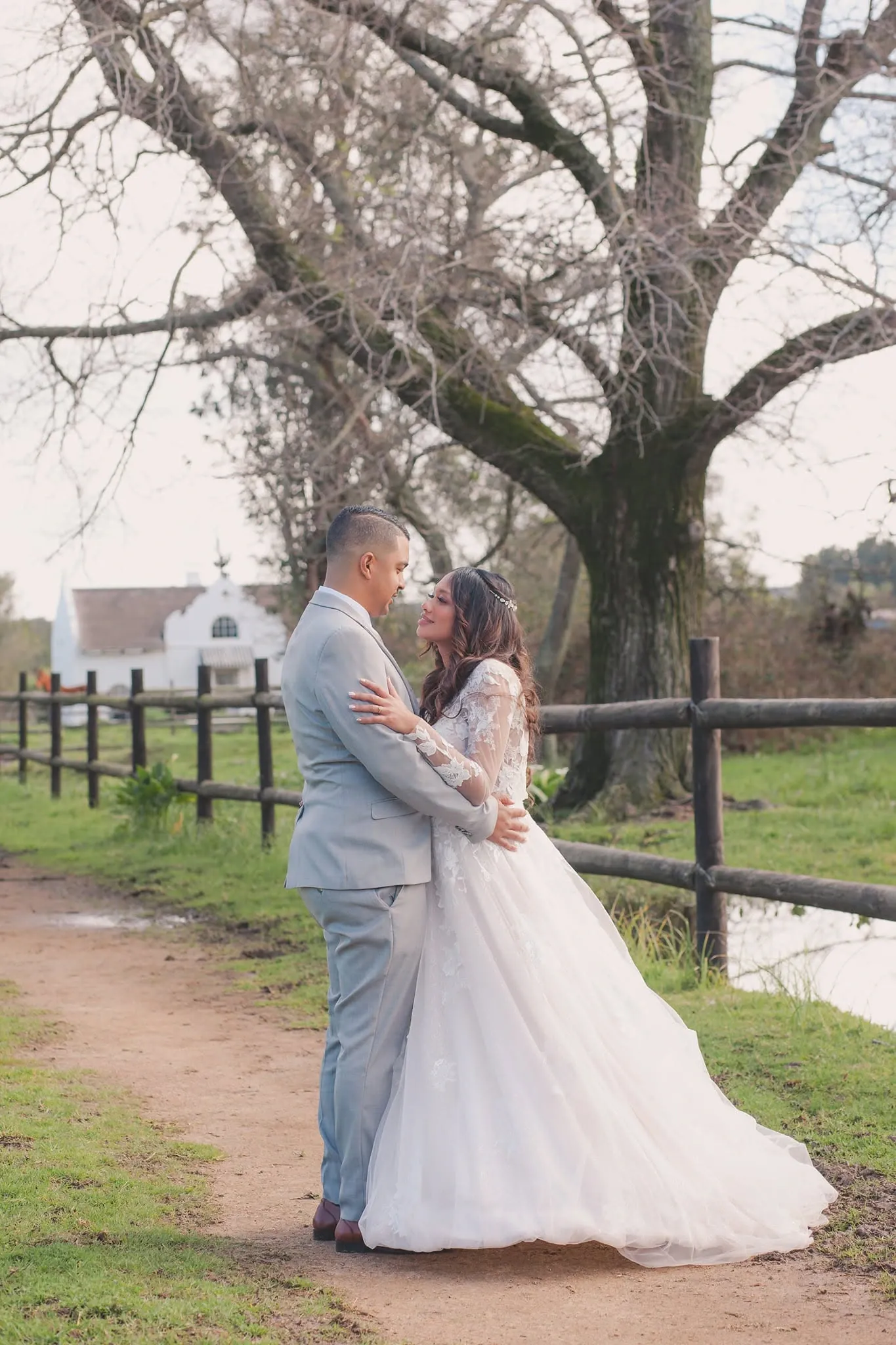 Bride in lace sleeves and groom in a pale grey suit embracing on a dirt path beside a wooden fence and a large bare oak, a white gabled building behind.