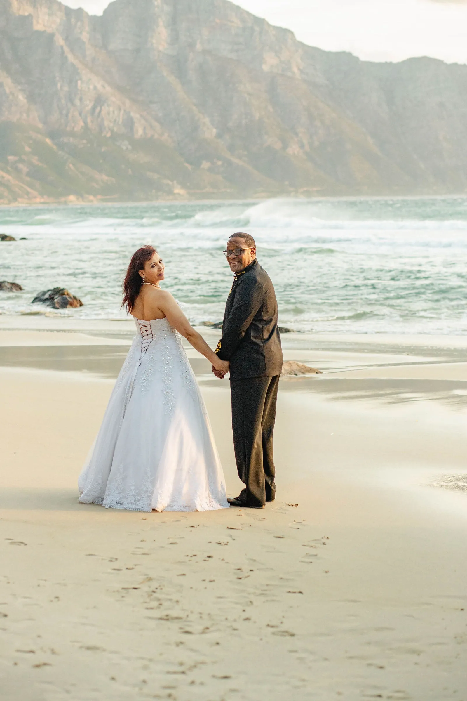 Bride and groom walking hand in hand on the beach towards the surf, both looking back at the camera, mountains behind the bay.