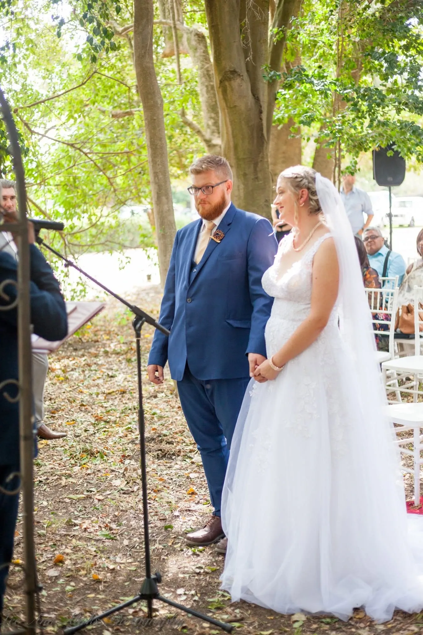 Bride and groom in a blue suit standing hand in hand before the officiant at an outdoor ceremony under tall trees.
