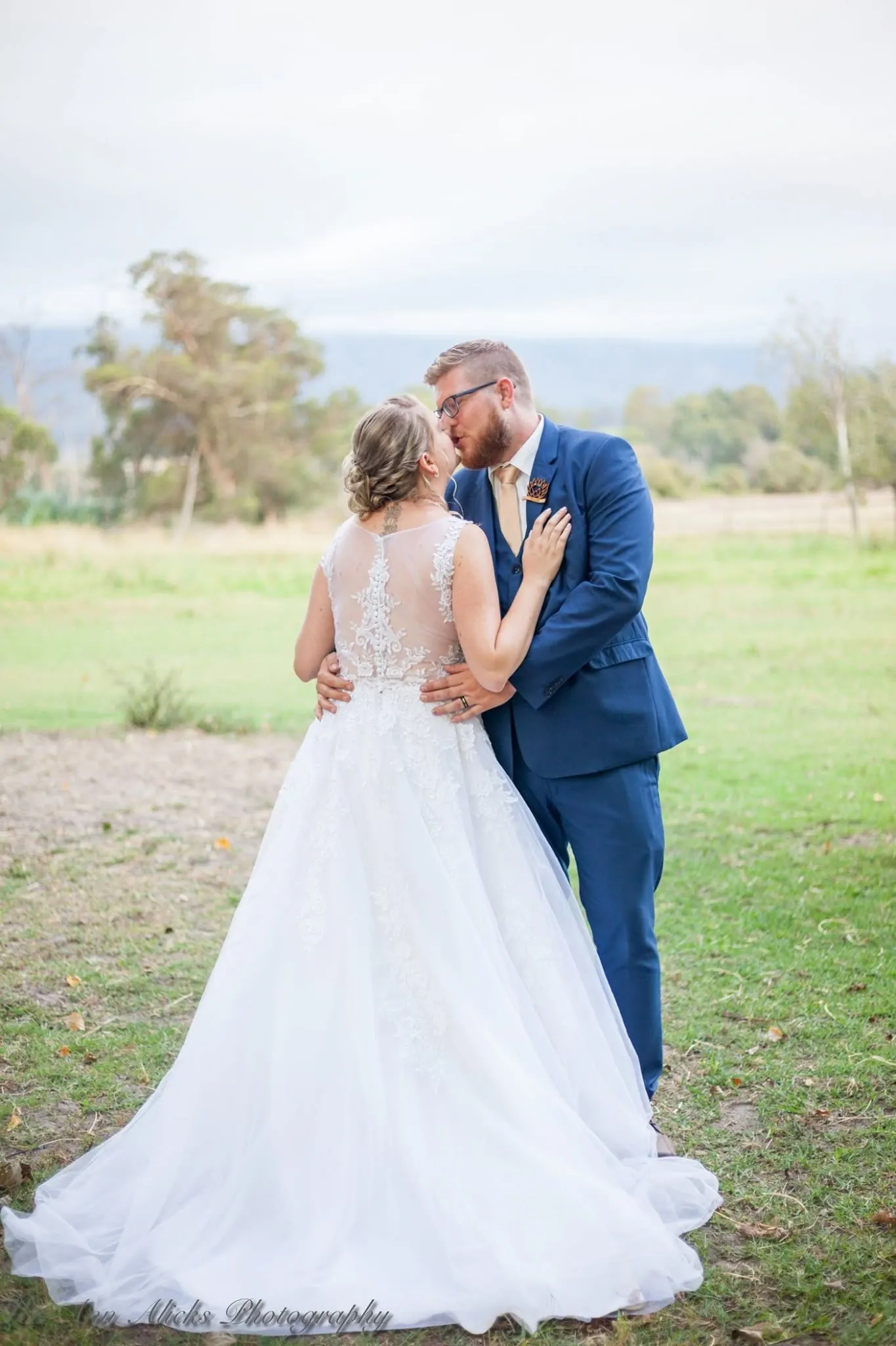 Bride and groom kissing in an open field, the bride's sheer lace back and full tulle skirt facing the camera.