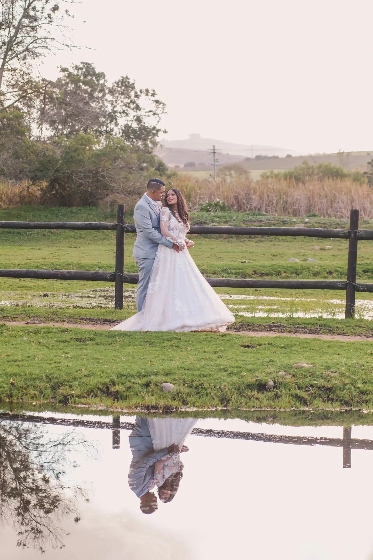 Bride and groom embracing by a wooden fence, the scene reflected upside down in still water in the foreground.