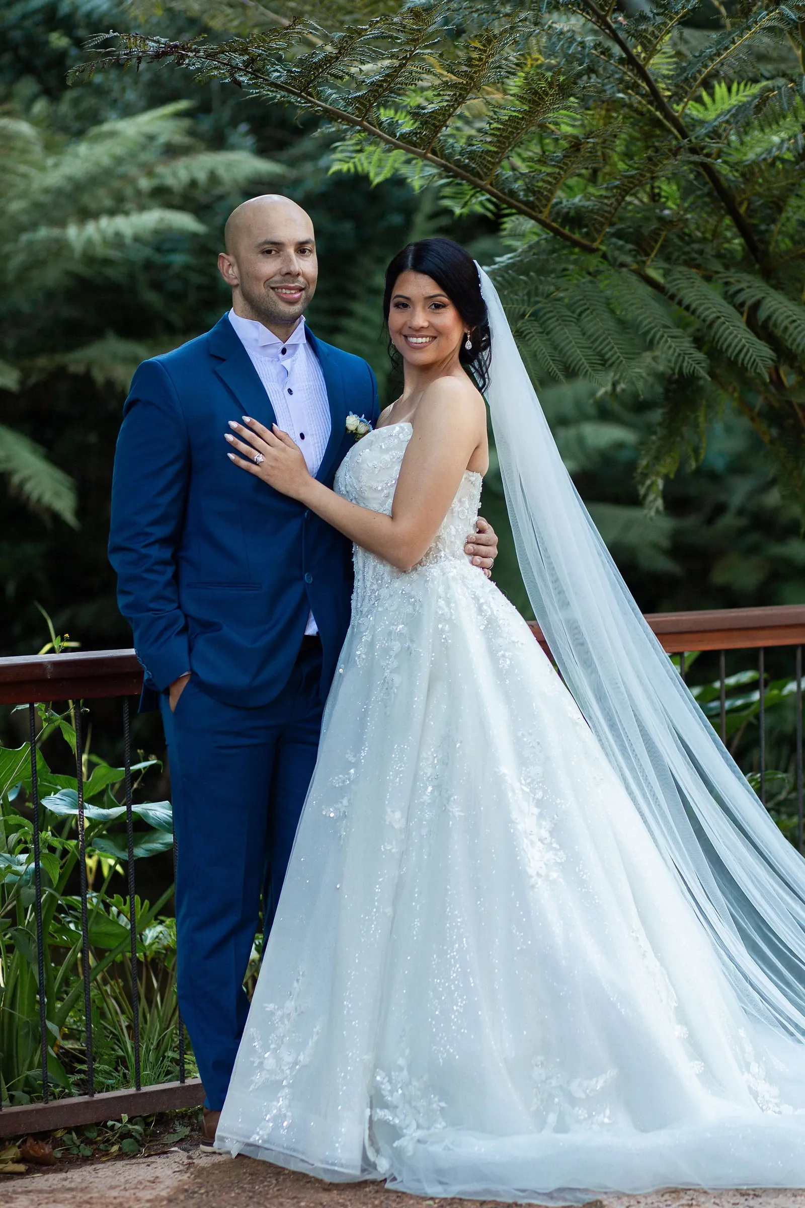 Groom in a royal-blue suit and bride in a beaded tulle gown with a long veil, standing in front of tree ferns.