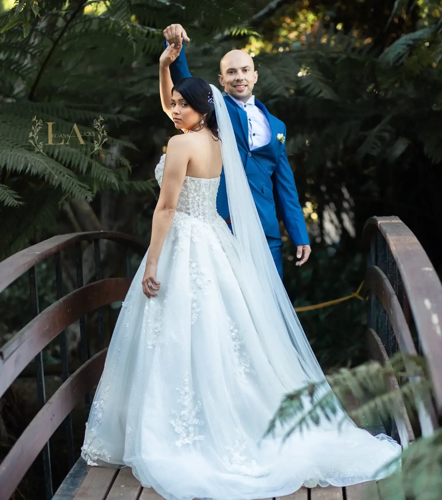 Groom twirling his bride on a wooden garden bridge, her veil and train trailing behind.
