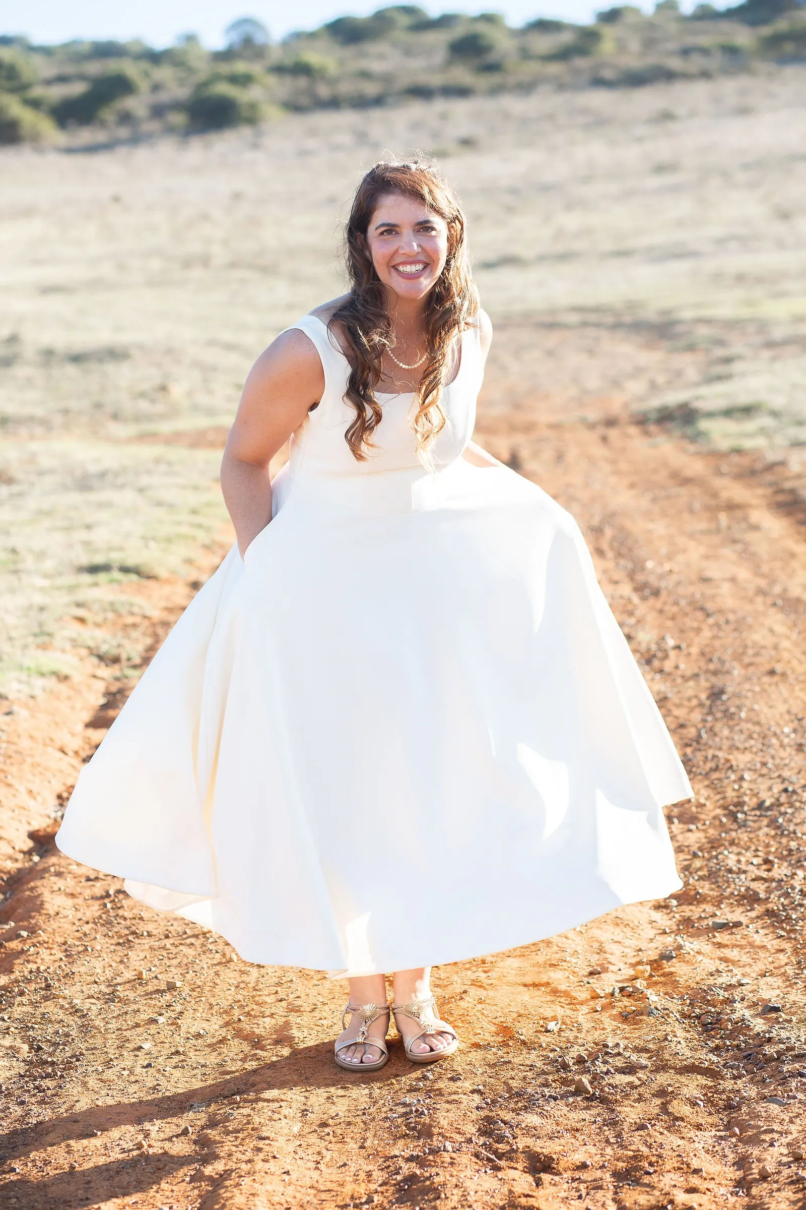 Bride in a satin tea-length dress holding out her skirt and grinning at the camera on a sunlit dirt track.