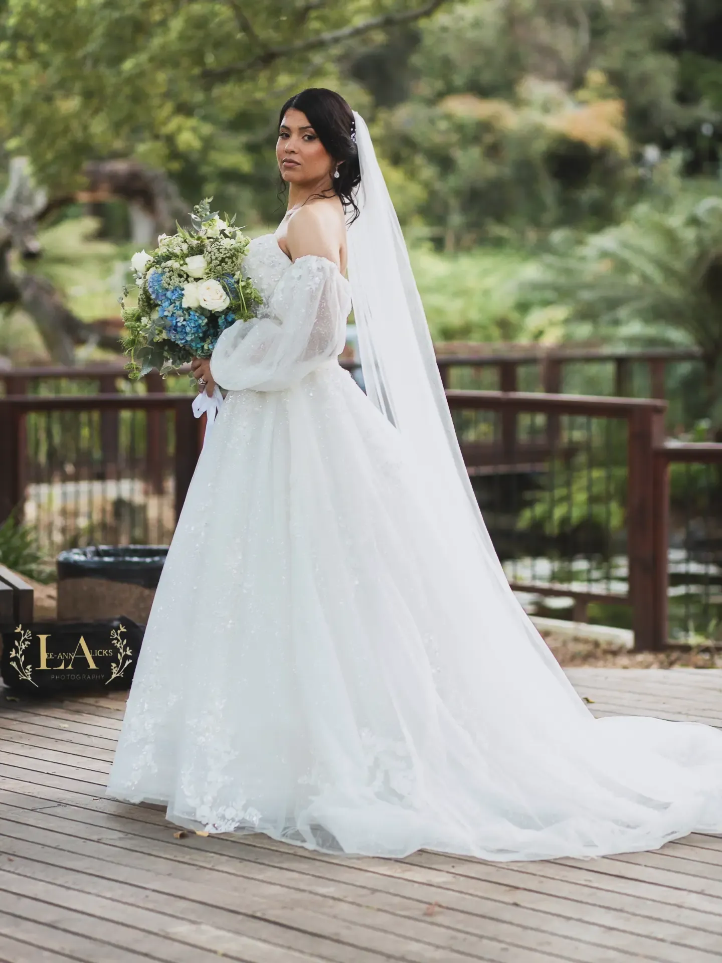 Bride in a lace gown with long sheer sleeves holding a blue and white bouquet on a garden deck.
