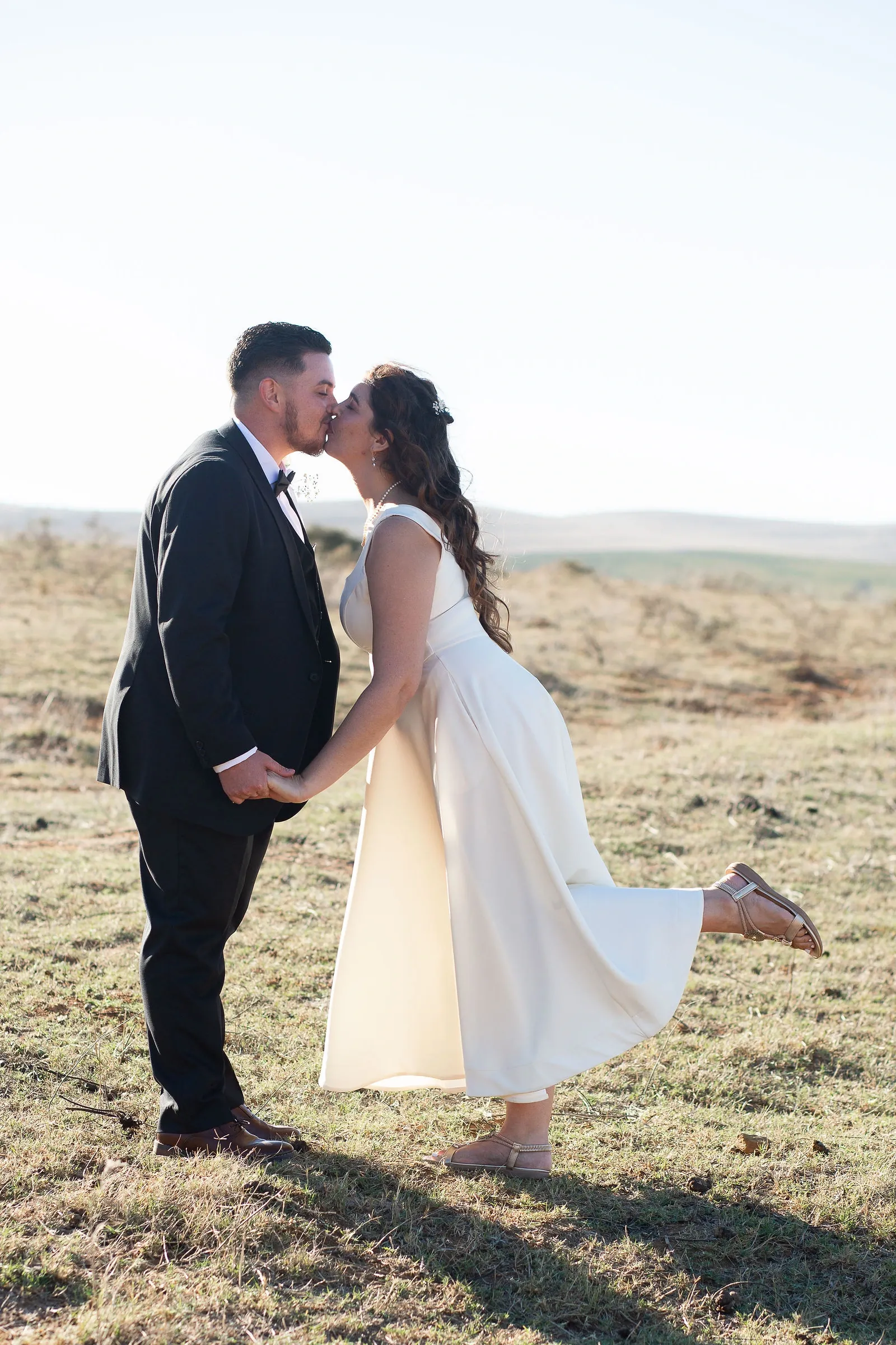 Backlit bride kissing her groom in a dry field, one foot kicked up behind her.
