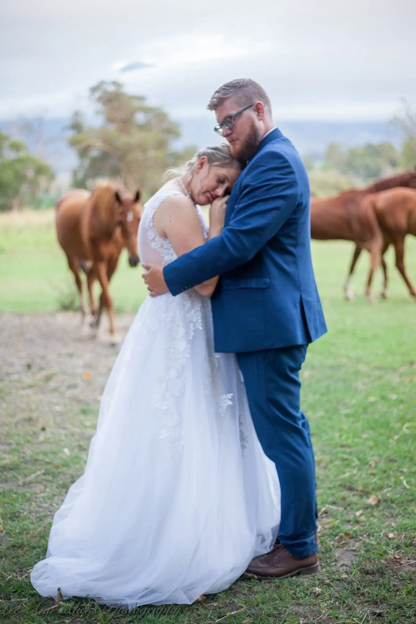 Bride resting her head on her groom's chest with her eyes closed while chestnut horses graze behind them.