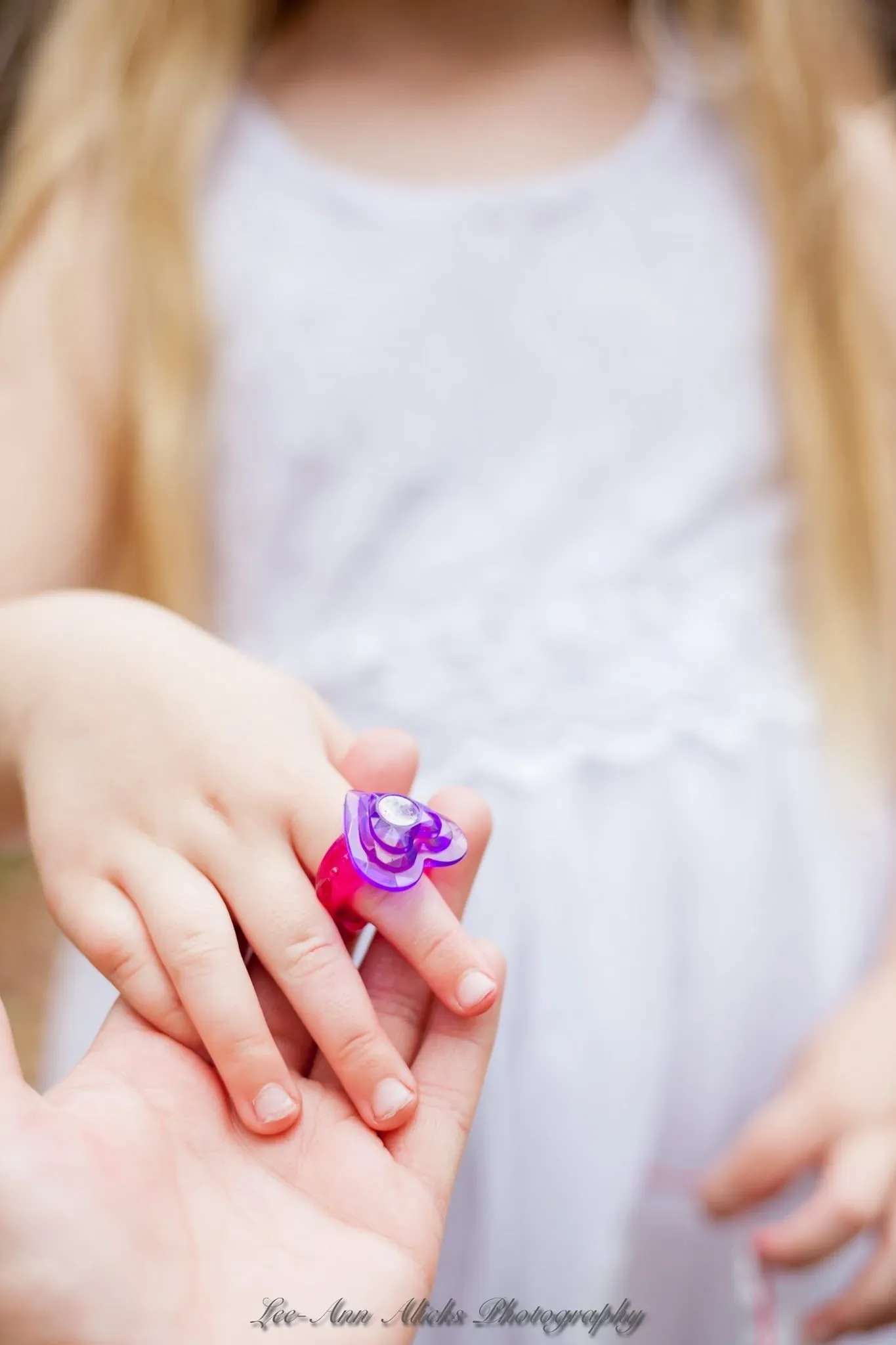 Close-up of a little girl's hand wearing a purple plastic heart ring, resting in an adult's open palm against her white dress.