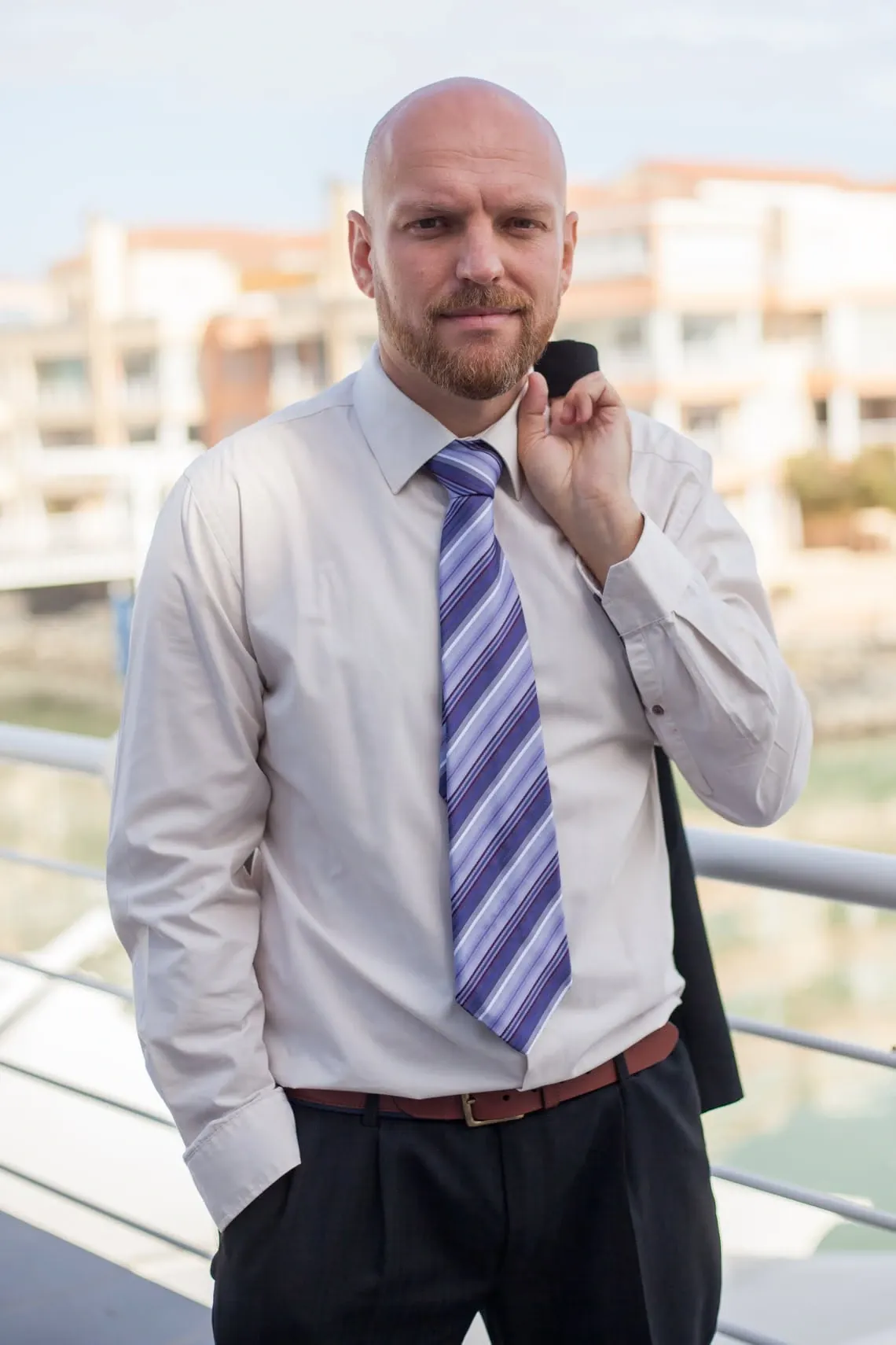 Corporate portrait of a man in a white shirt and striped tie, jacket over his shoulder, outdoors by the water.