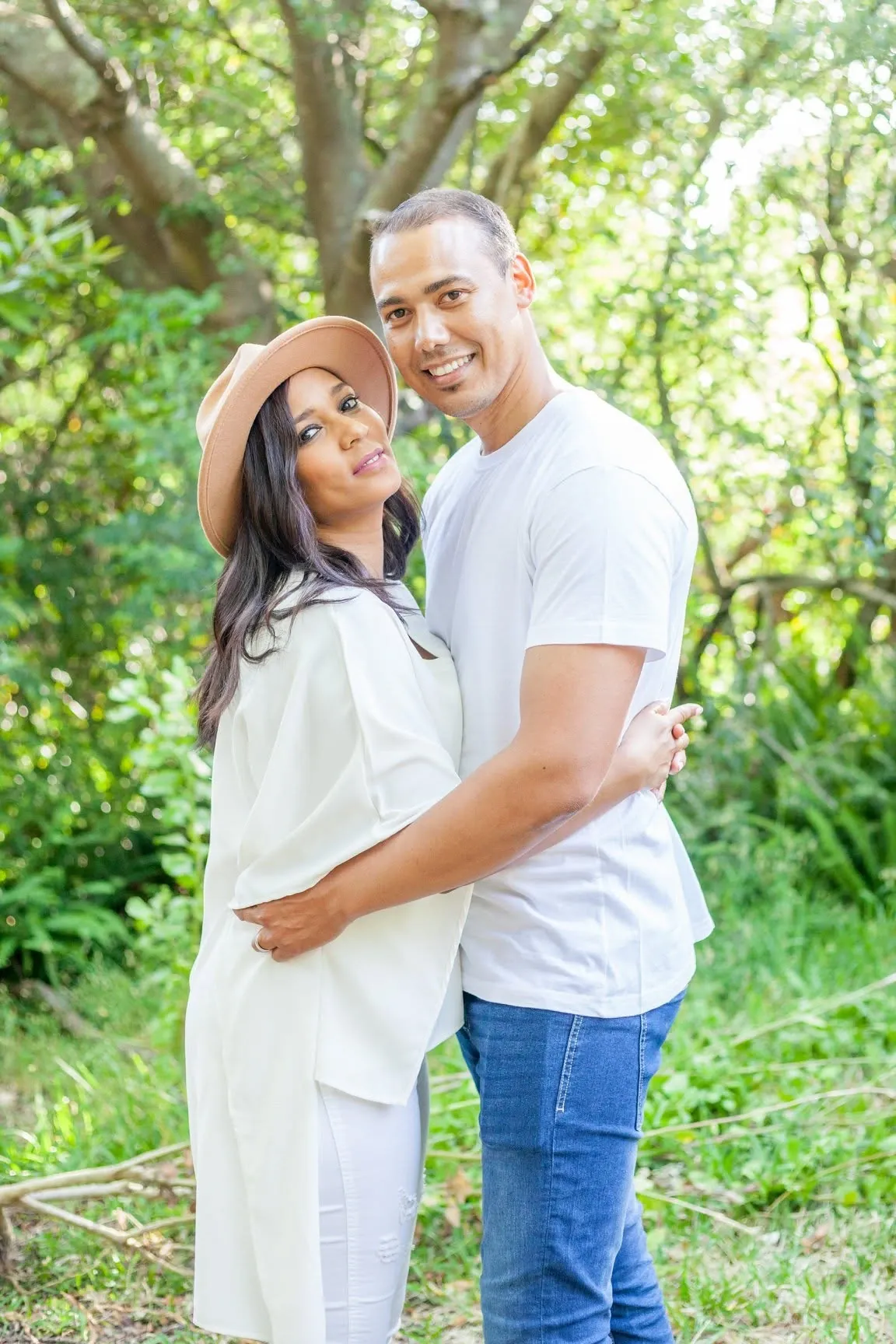Couple in white tops hugging among green trees, she in a tan fedora glancing back over her shoulder at the camera.