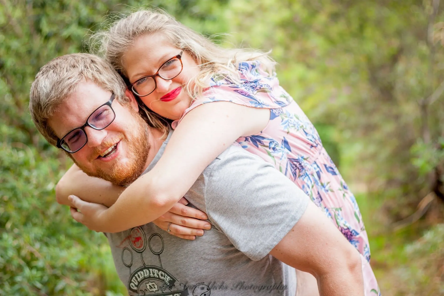 Woman in glasses and red lipstick riding piggyback on her grinning partner among green shrubs.