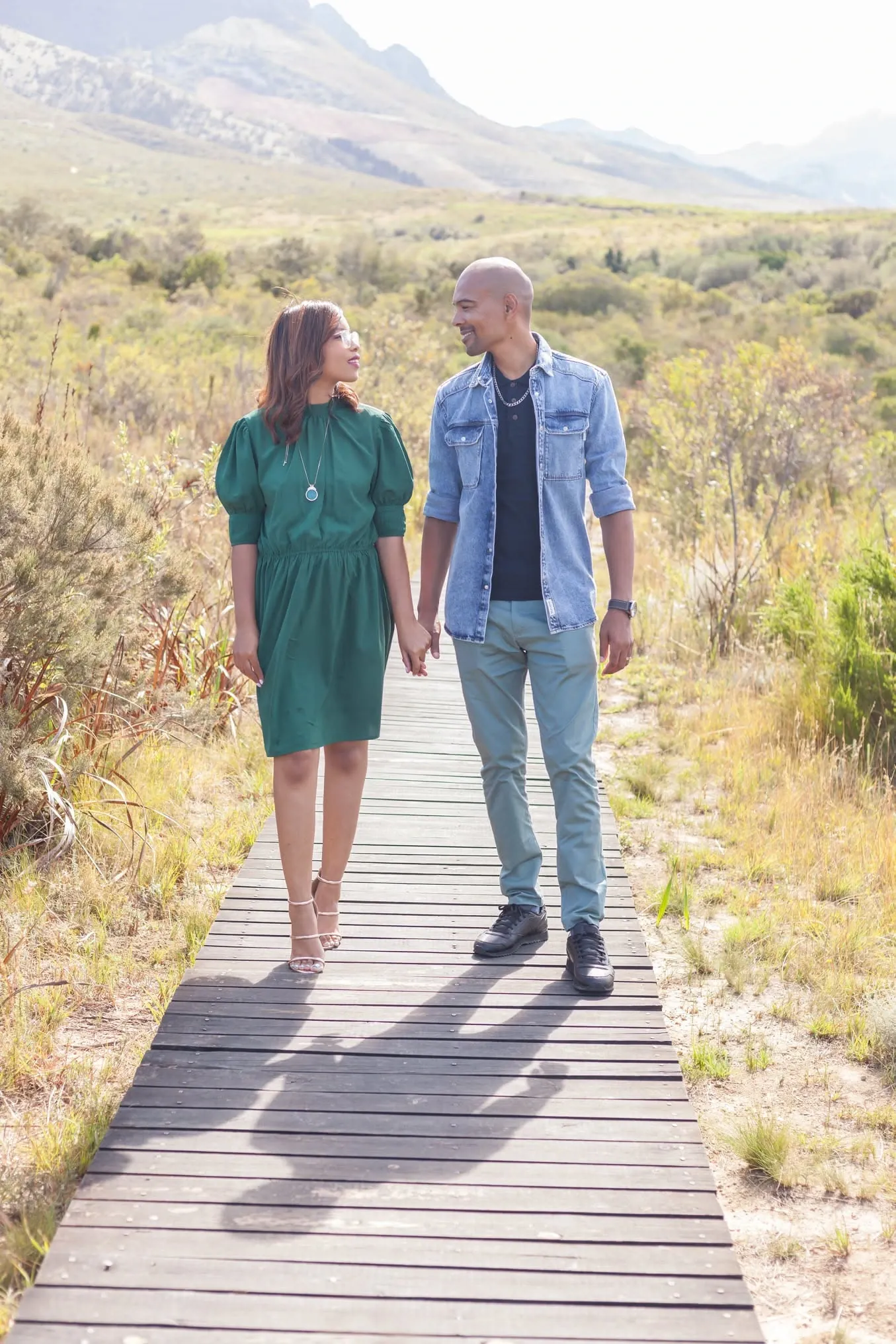 Couple walking hand in hand along a wooden boardwalk through fynbos, smiling at each other.