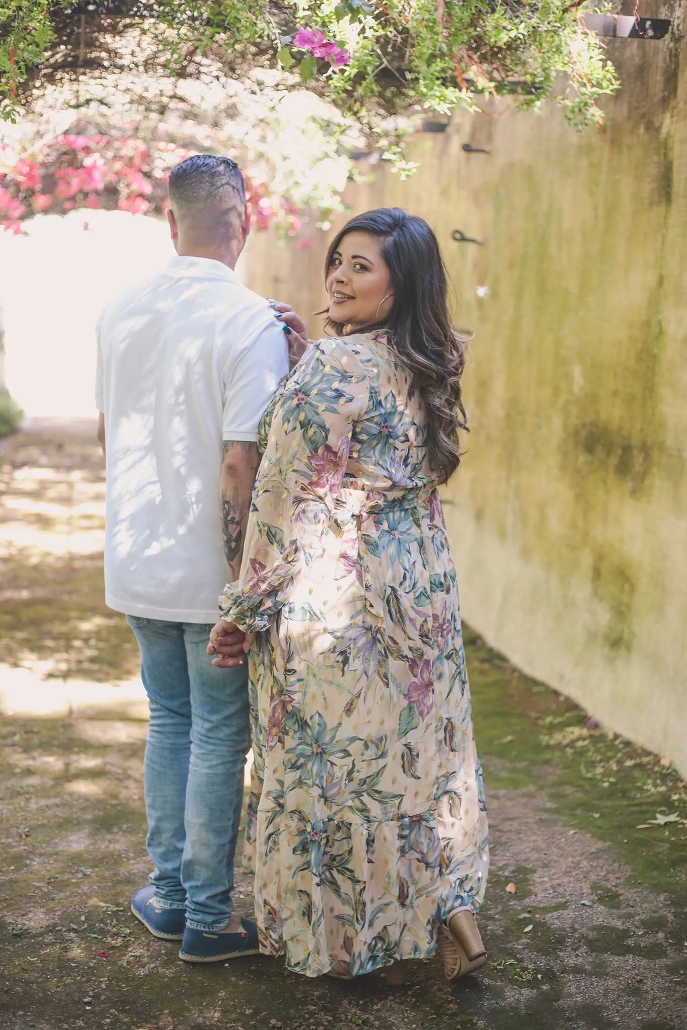 Couple walking away down a garden lane beside a mossy wall, she looks back over her shoulder in a floral maxi dress.