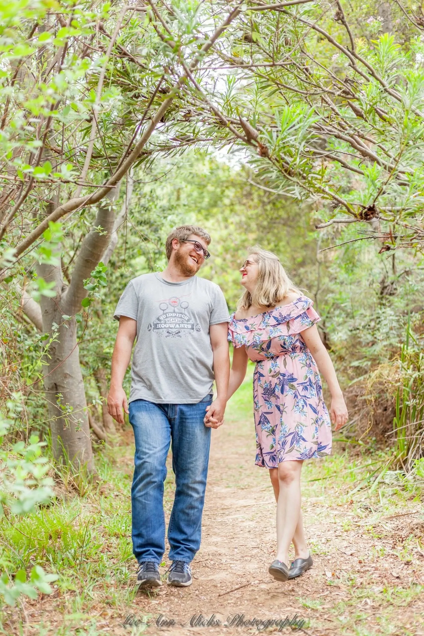 Couple laughing at each other as they walk hand in hand down a dirt trail under arching branches.