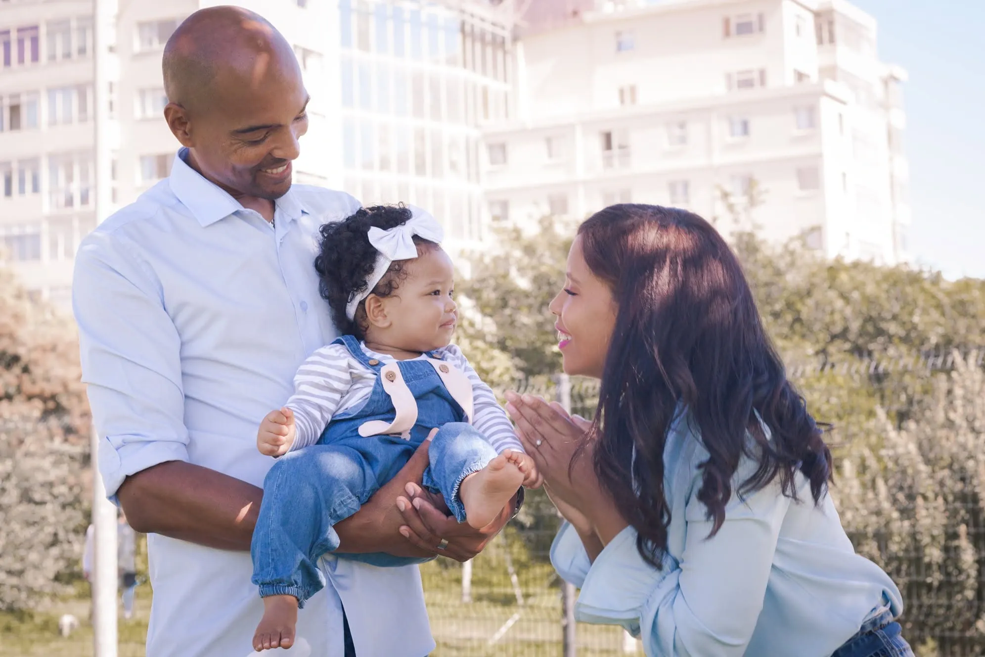Dad holding his baby daughter in denim dungarees while mum claps and smiles at her.