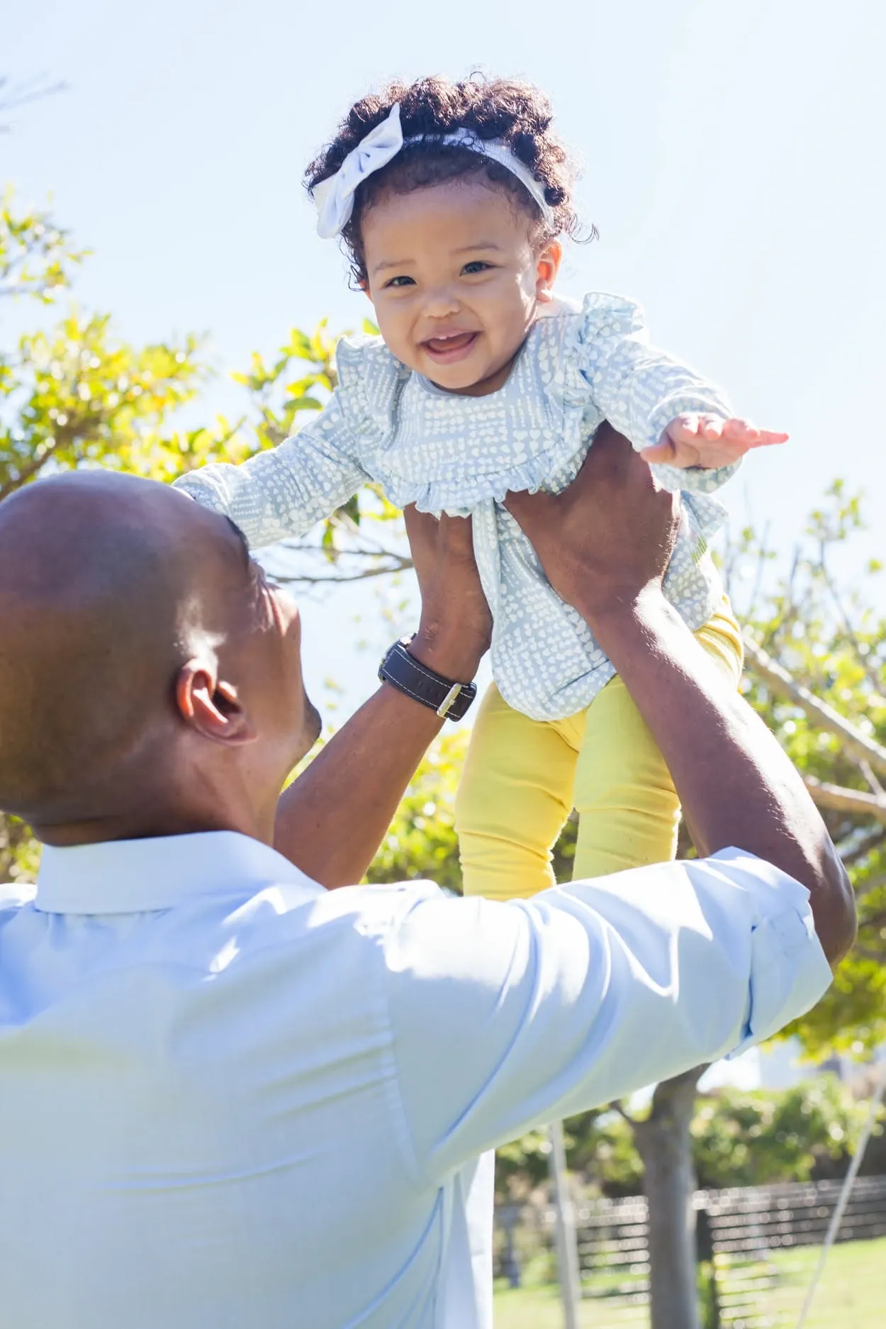 Dad lifting his smiling baby daughter overhead against a clear sky.