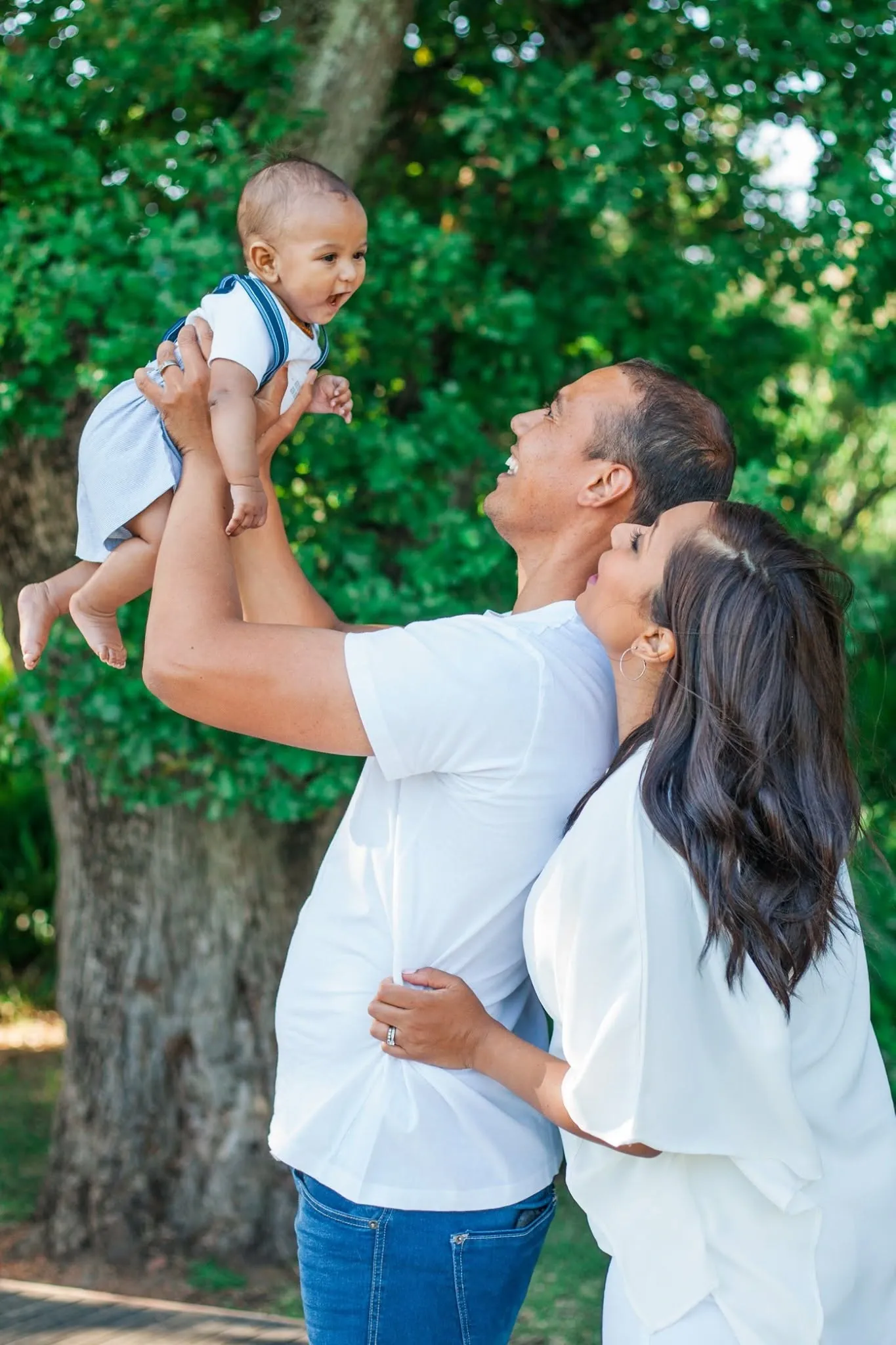 Dad holding his baby son up high under a big tree while mum holds him around the waist, both laughing.