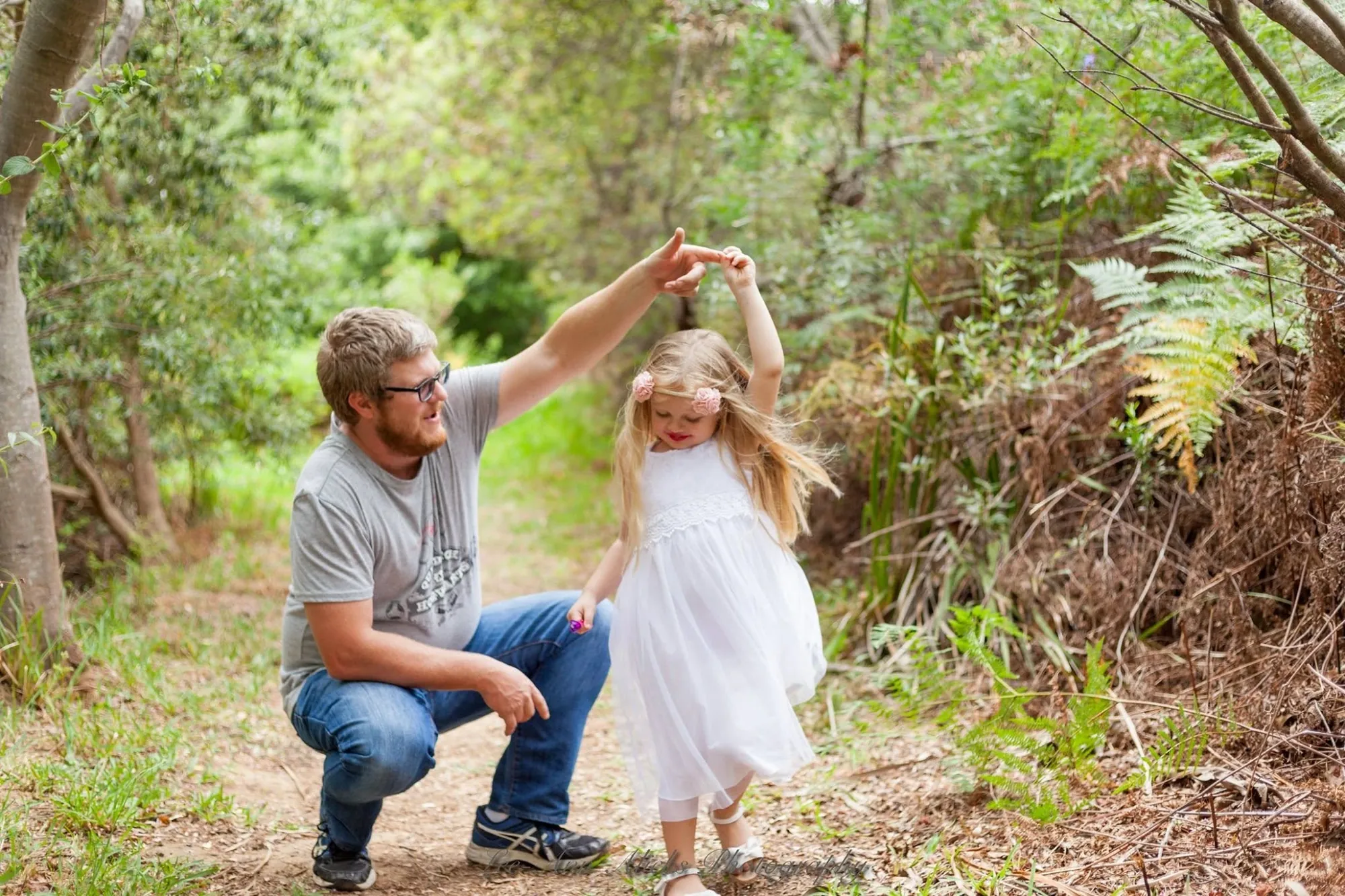 Dad crouching on a woodland path as he twirls his small daughter in a white dress by the hand.