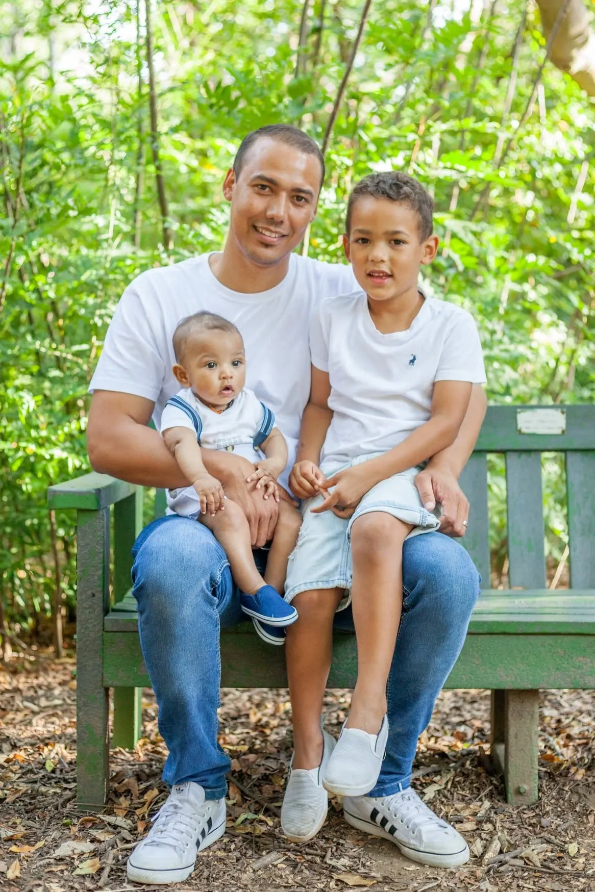 Dad seated on a green wooden garden bench with his baby son and older son on his knees, all in white T-shirts.