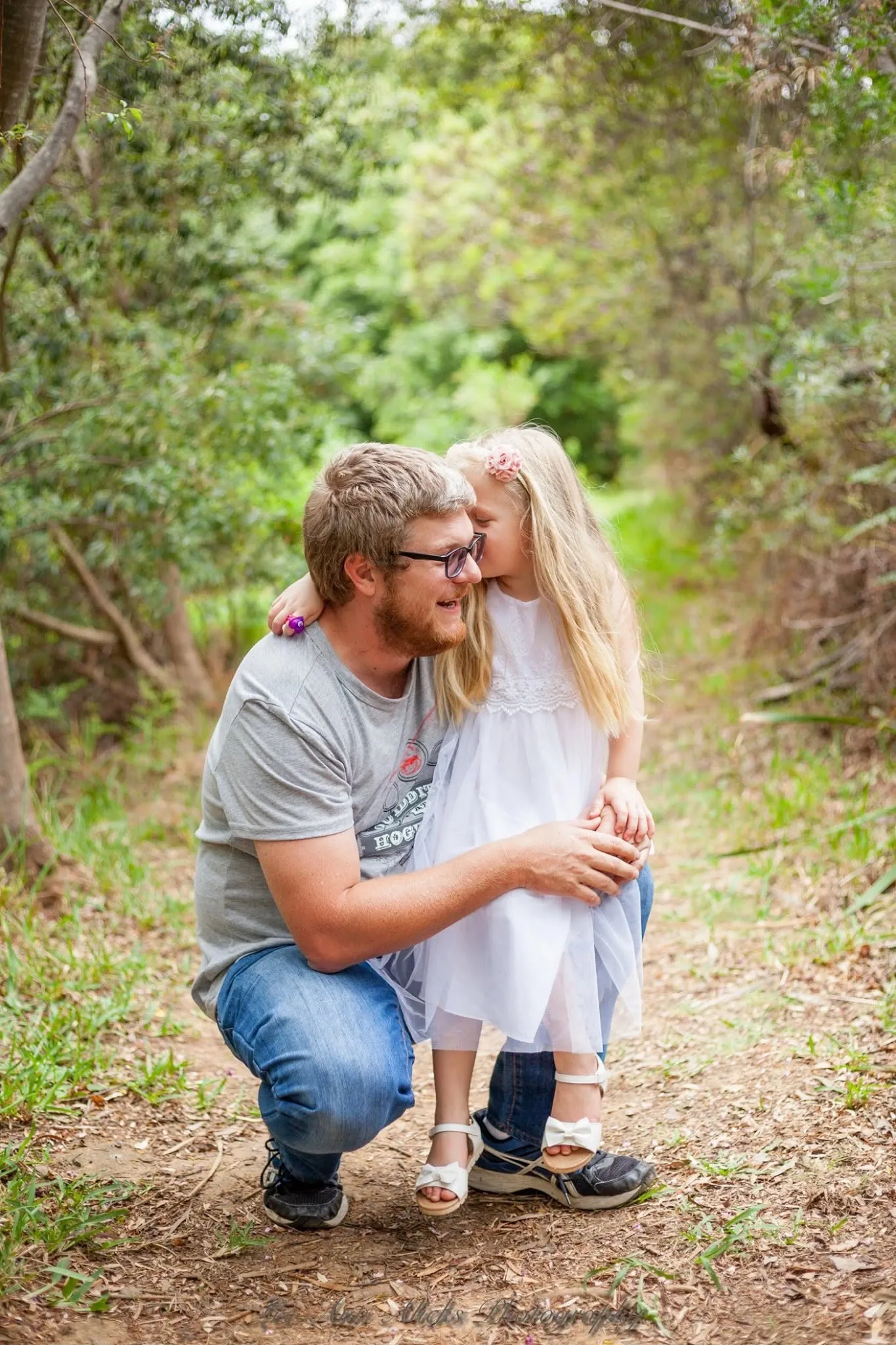 Little girl in a white tulle dress kissing her crouching dad on the cheek on a forest path.