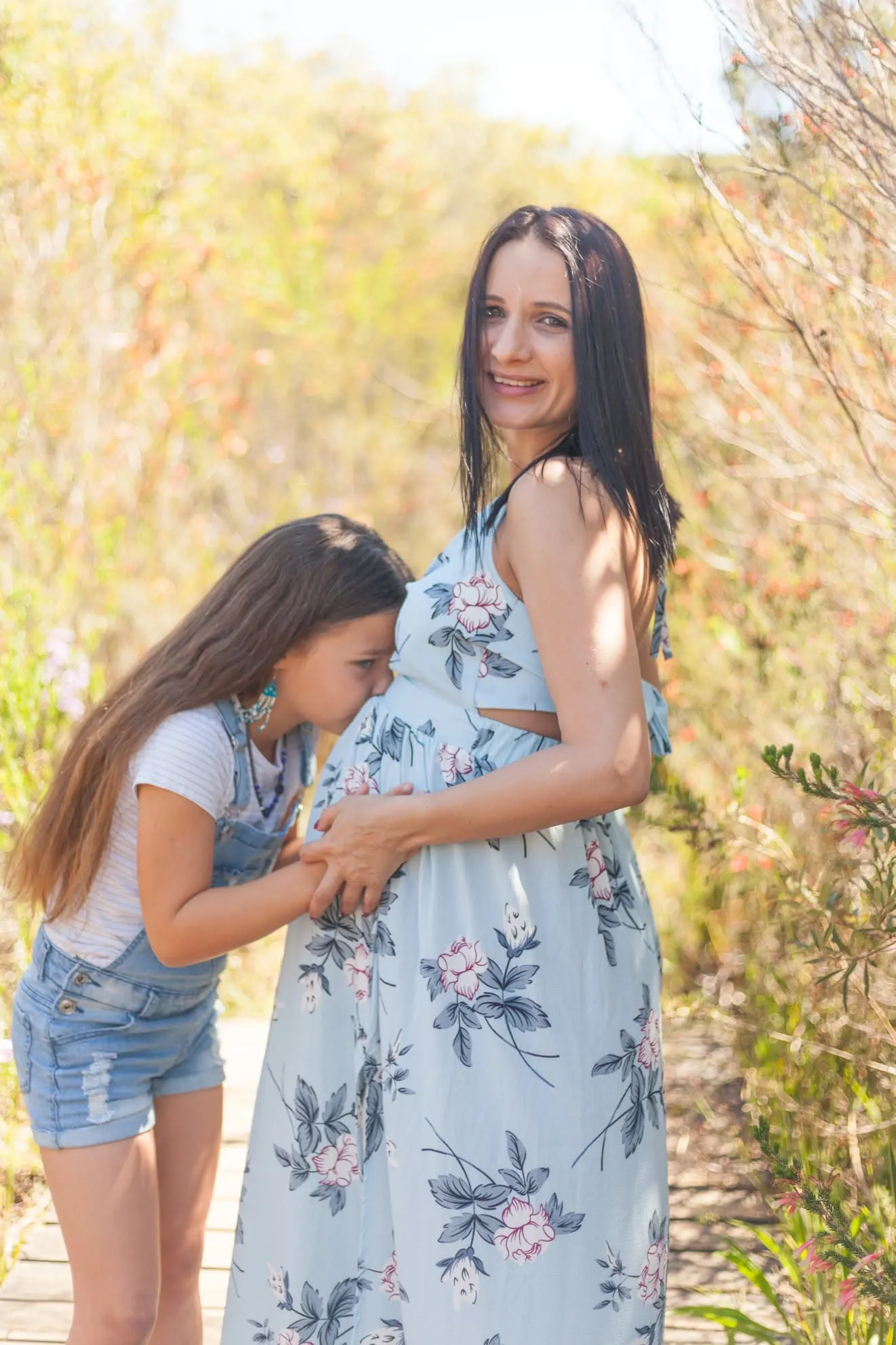 Girl in denim dungarees kissing her mother's bump on a boardwalk while mum smiles at the camera.