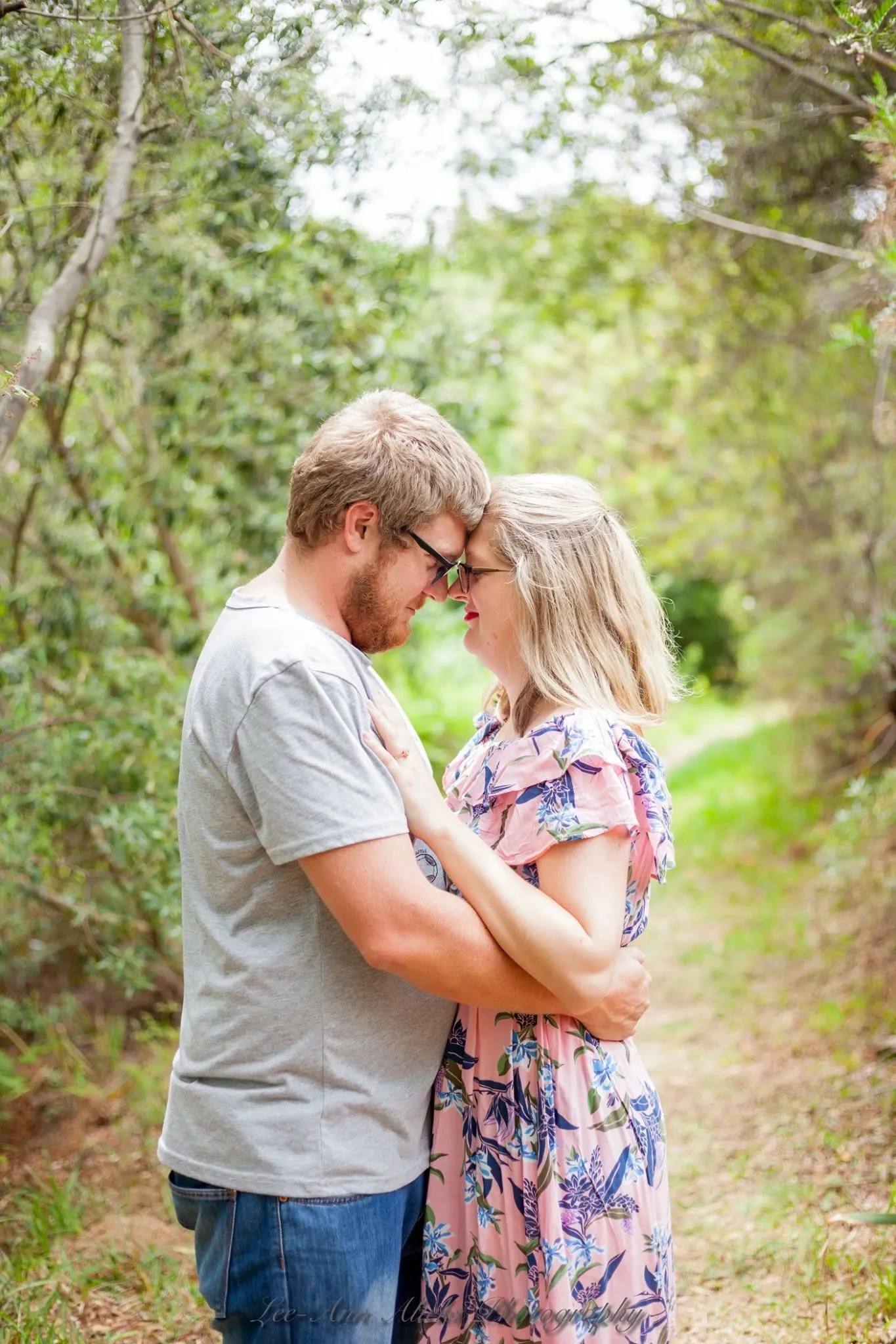 Couple standing forehead to forehead on a leafy forest path, he in a grey T-shirt and glasses, she in a pink floral dress.