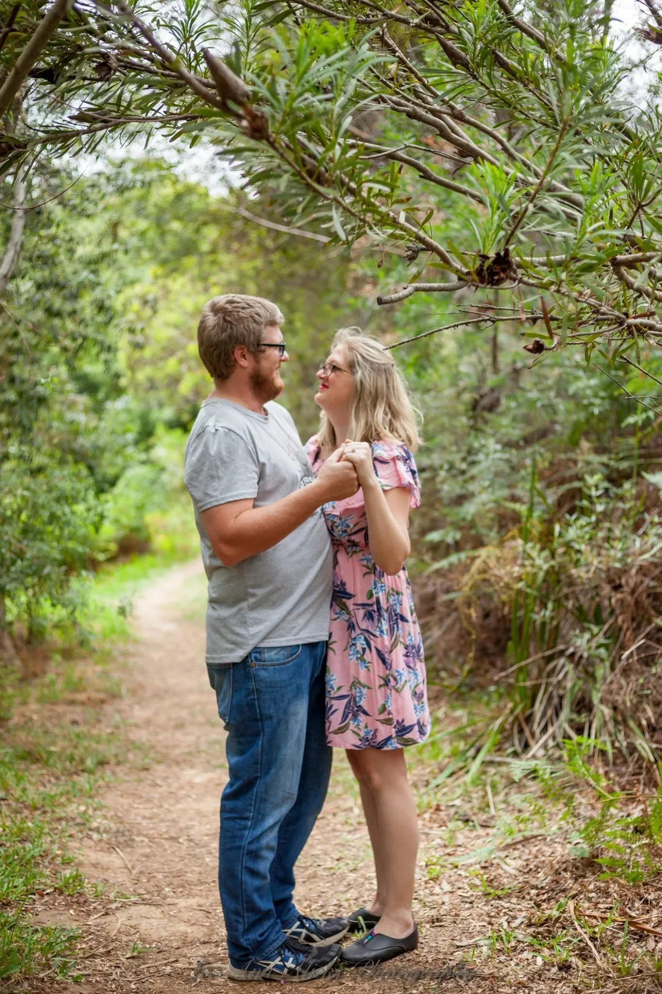 Couple holding hands at chest height and gazing at each other on a dirt path under overhanging branches.