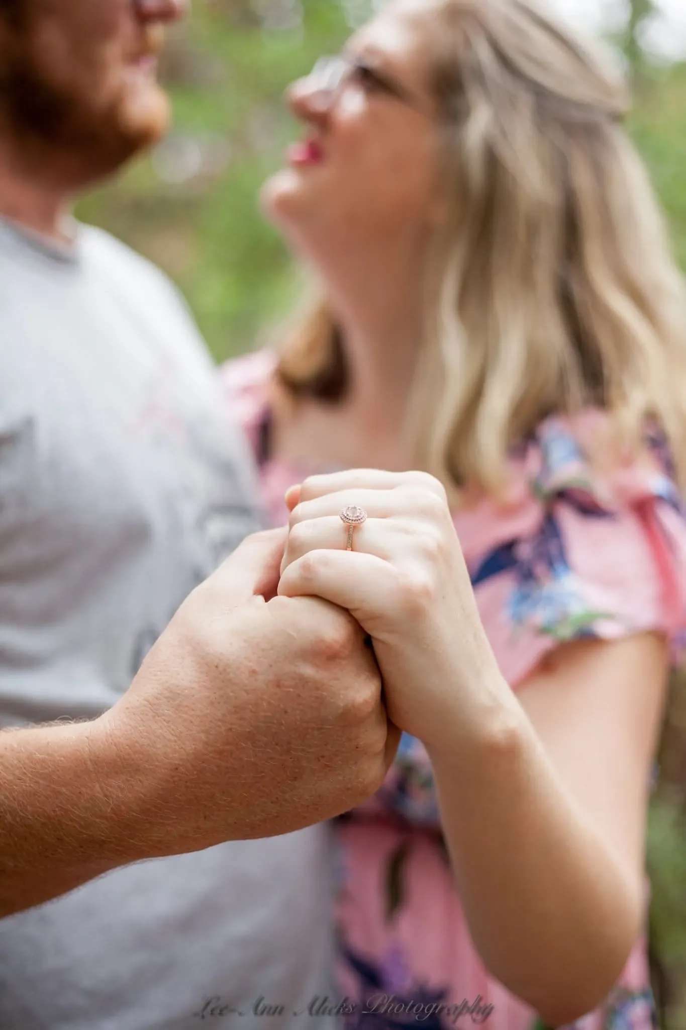 Close-up of a couple's clasped hands showing her rose-gold engagement ring, her laughing face soft in the background.