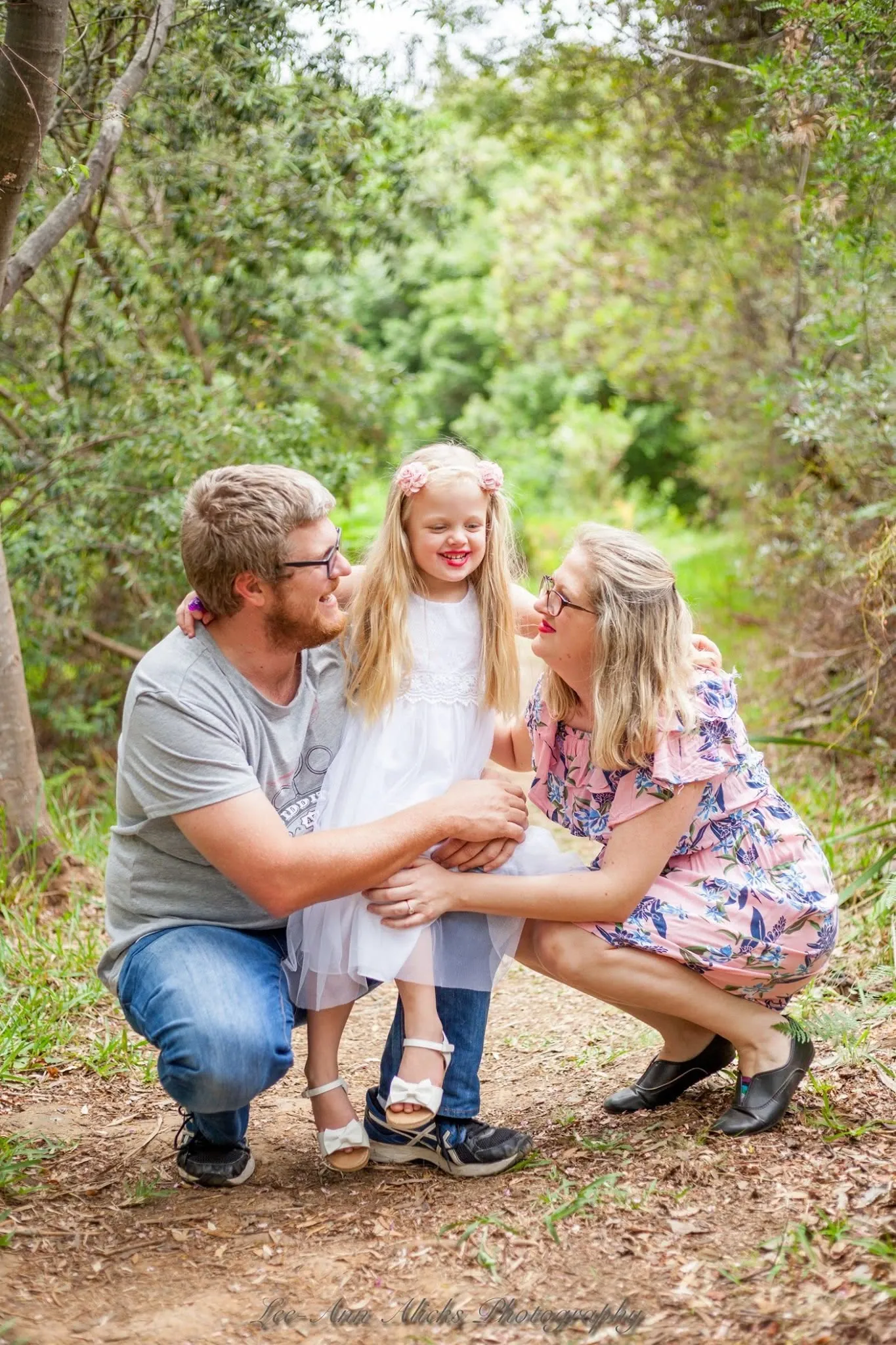 Couple crouching on a leafy forest path with their arms around a laughing little girl in a white tulle dress and pink flower clips.