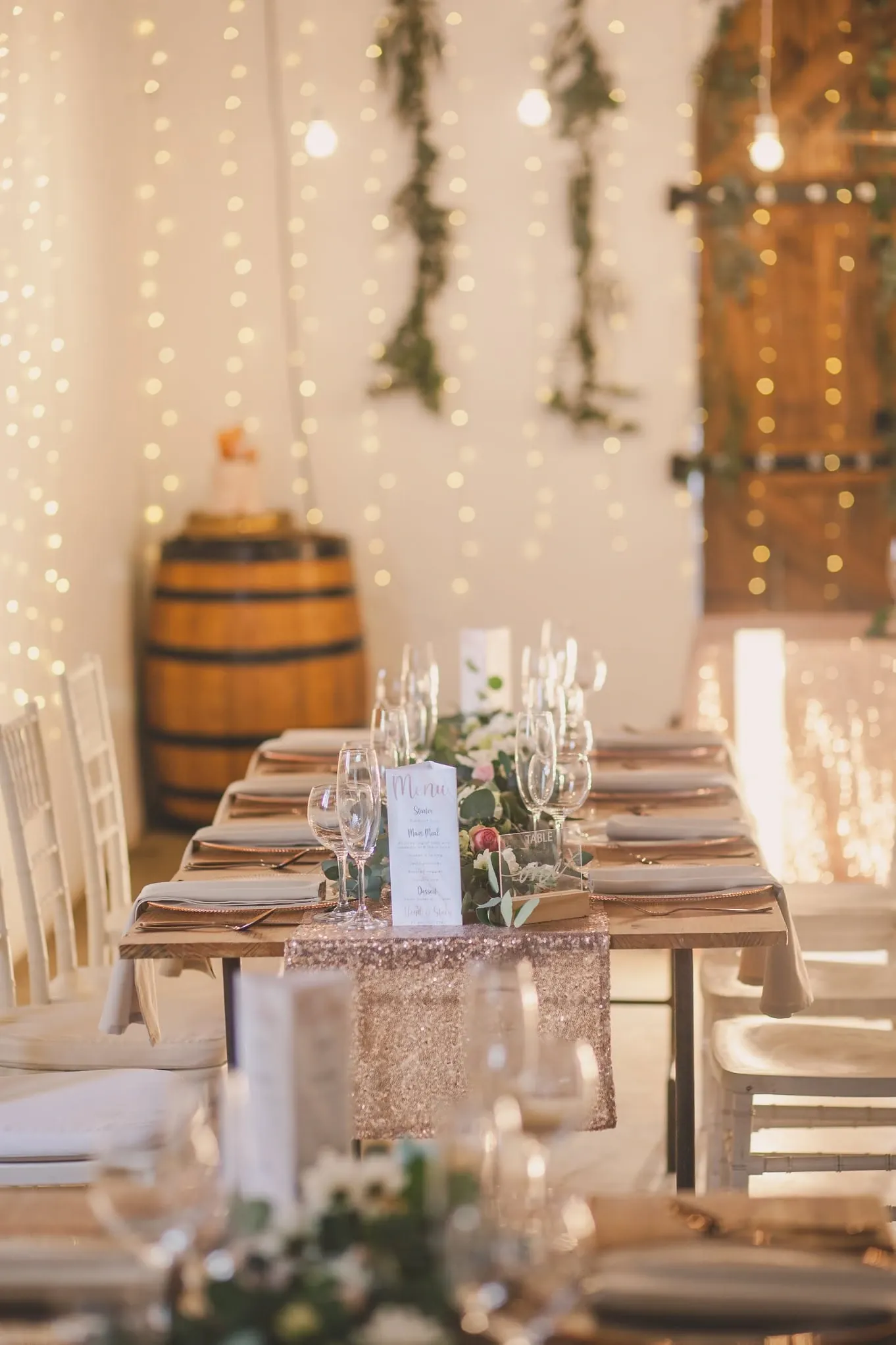 Venue set for a celebration: long wooden tables, glassware and greenery in front of fairy lights and a wine barrel.