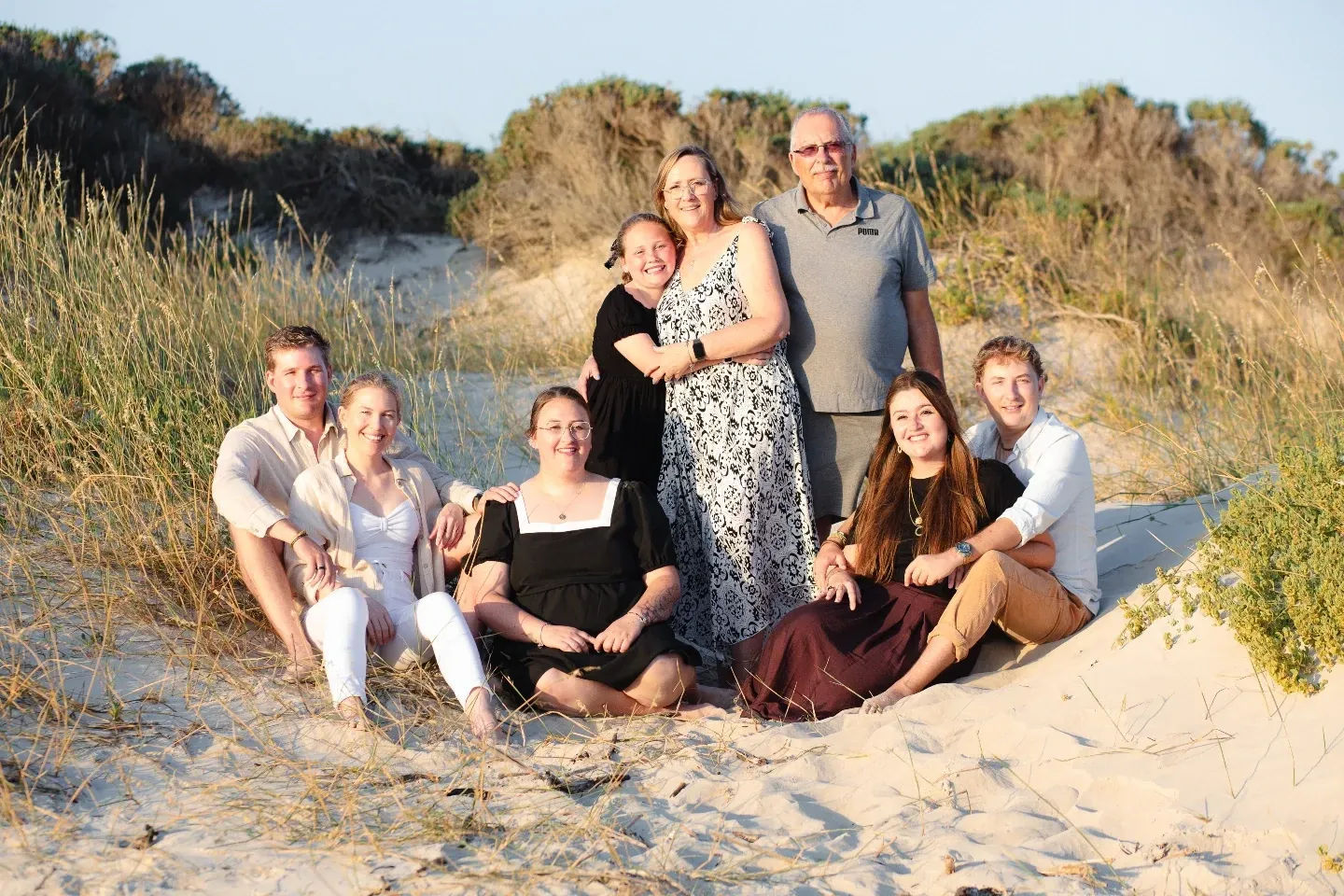 Extended family of eight seated and standing together among dune grass in late afternoon light.
