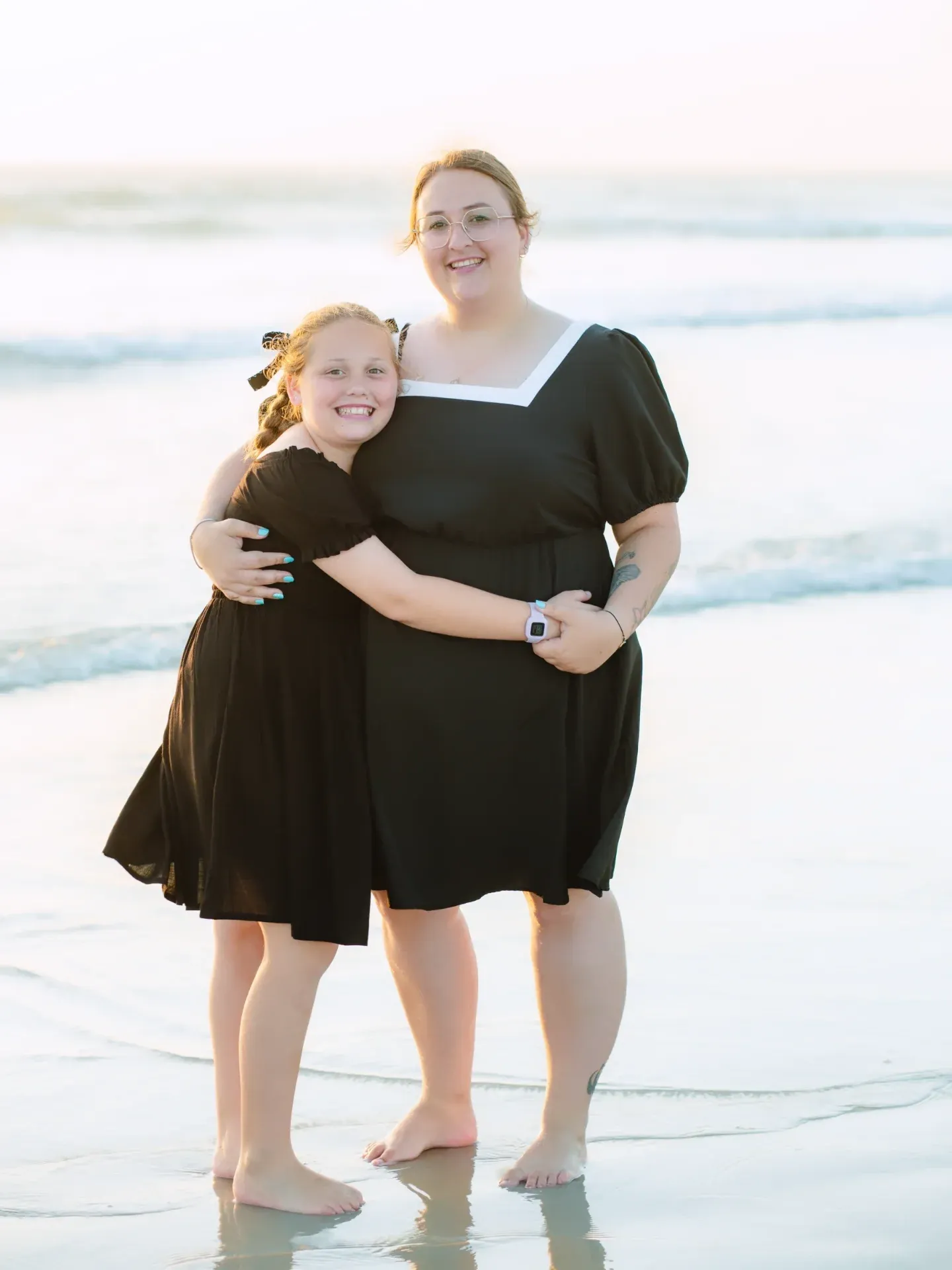 Mother and daughter hugging barefoot in the shallow water during a family beach photoshoot.