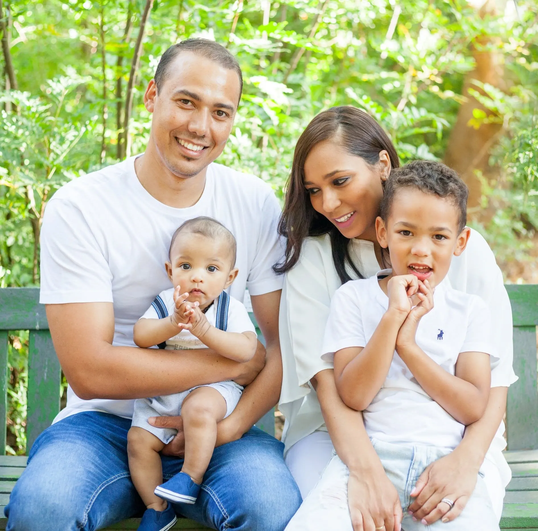 Family of four in white tops on a green garden bench, the baby wide-eyed and the older boy with his hands at his mouth.