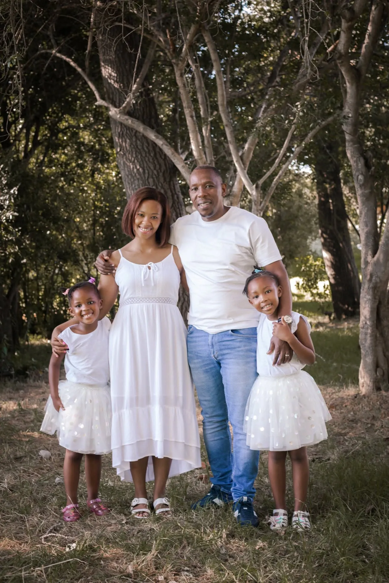 Family of four in white standing together under tall trees, the two girls in tulle skirts.
