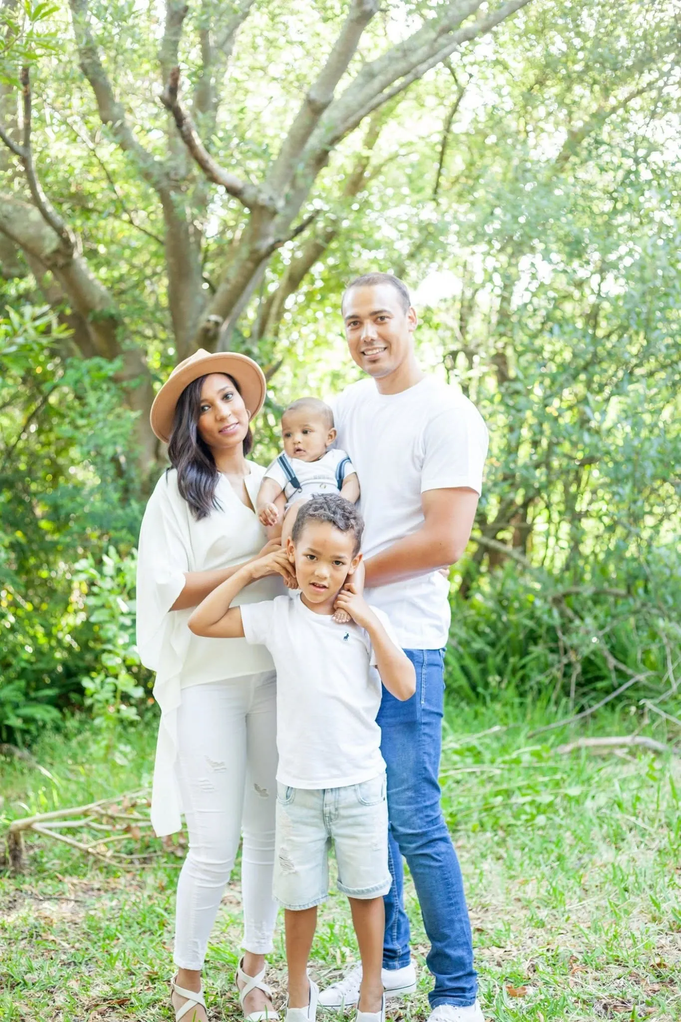 Family of four in white tops standing close together under a spreading tree, the baby held up between his parents.