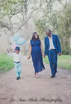Pregnant mum and dad walking hand in hand down a tree-lined path with their son, who holds pastel balloons.