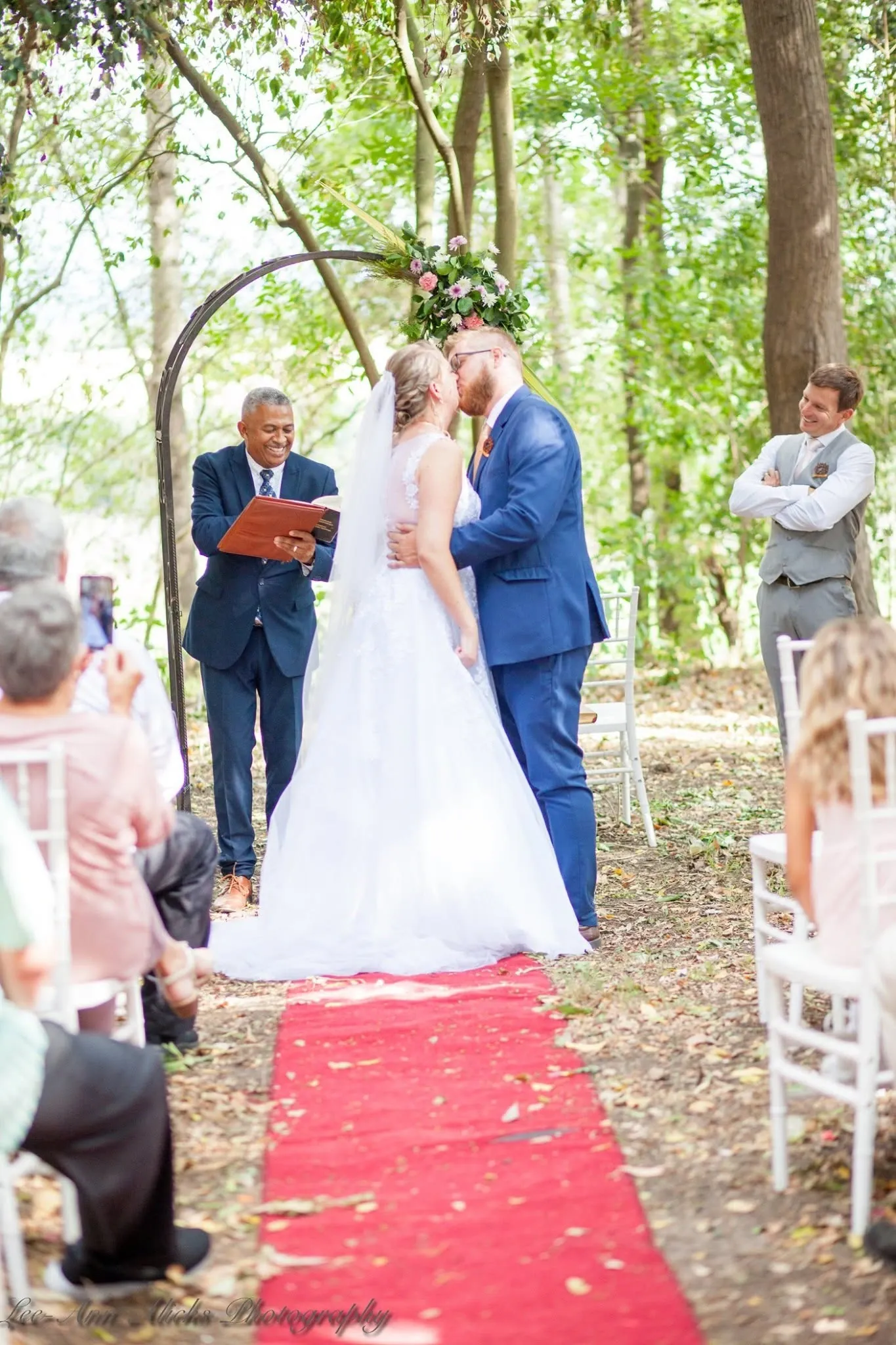 Bride and groom kissing under a flower-trimmed iron arch at the end of a red carpet aisle, the officiant smiling beside them.