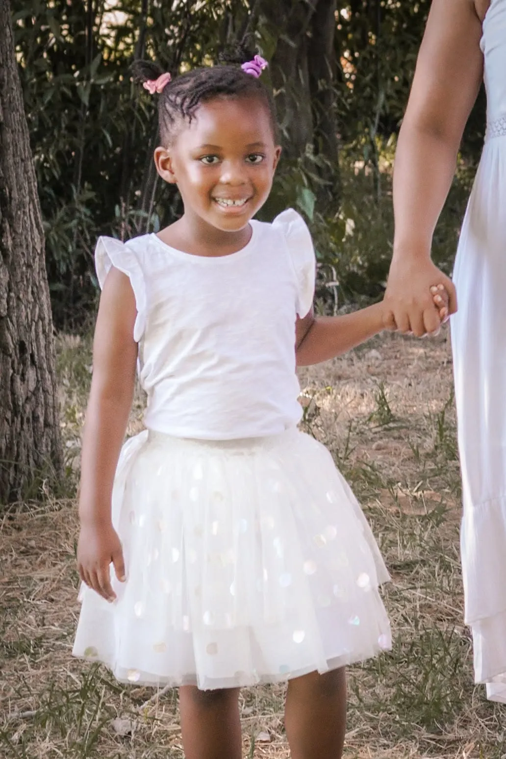 Smiling little girl in a white tulle skirt holding her mother's hand under trees.